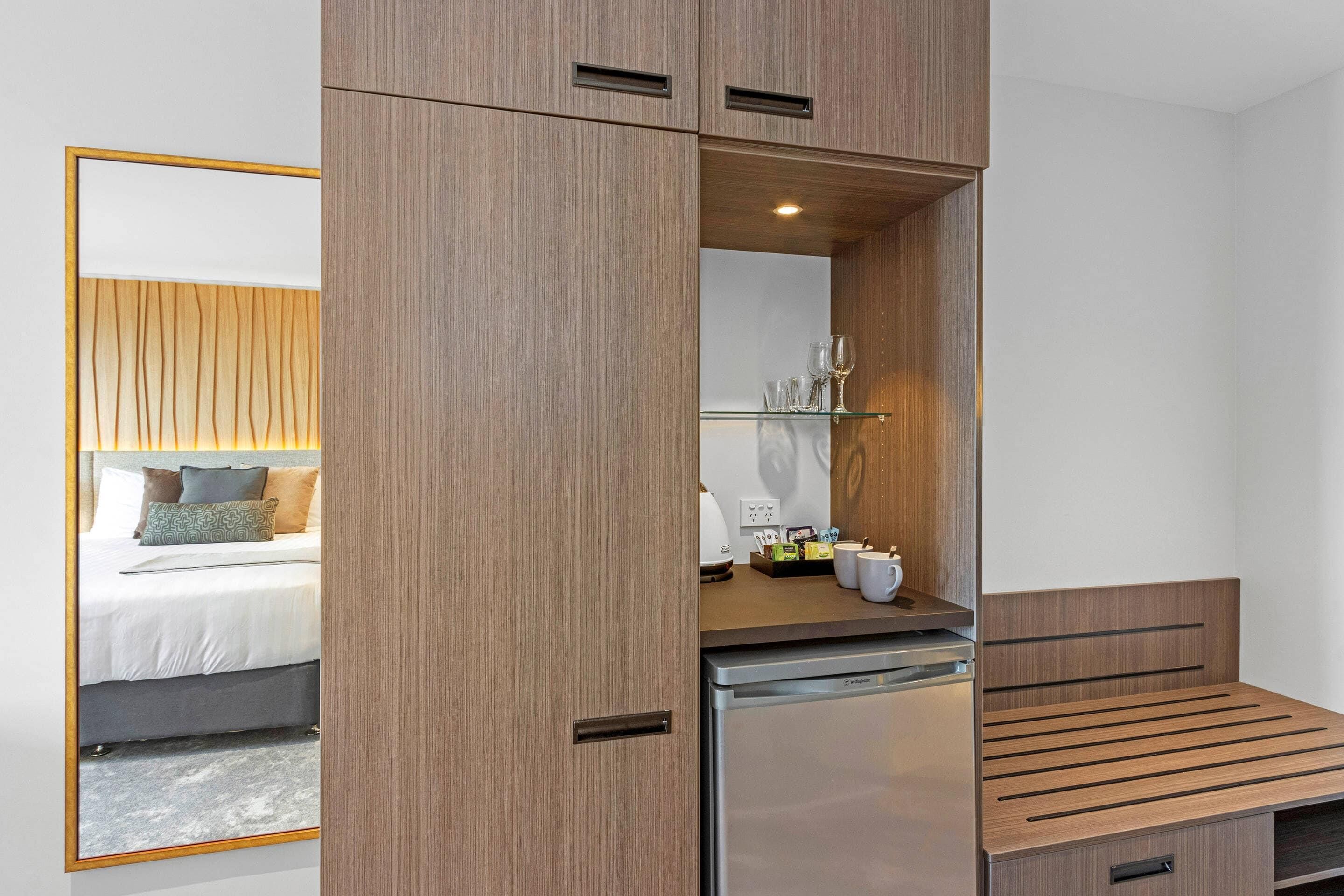 A compact room with a bed, a small kitchenette, a mirror, and a seating area.