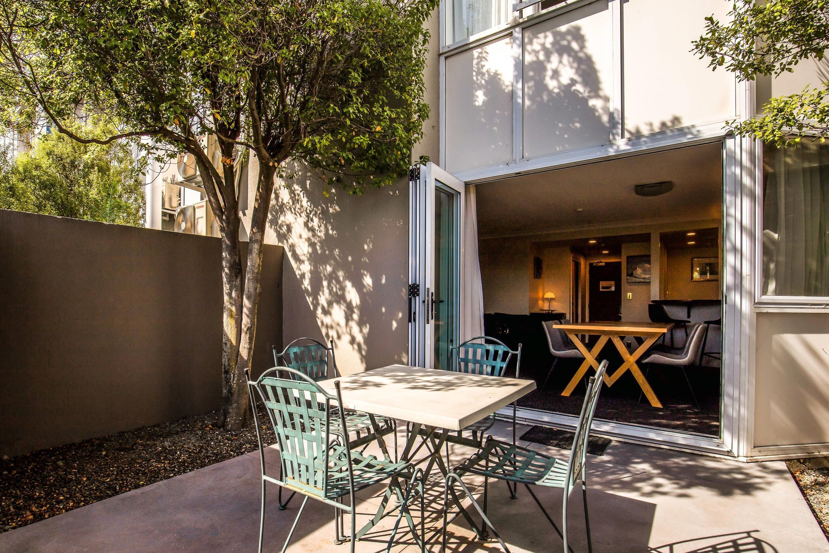 An open patio with a table and chairs, a tree, and a building entrance.