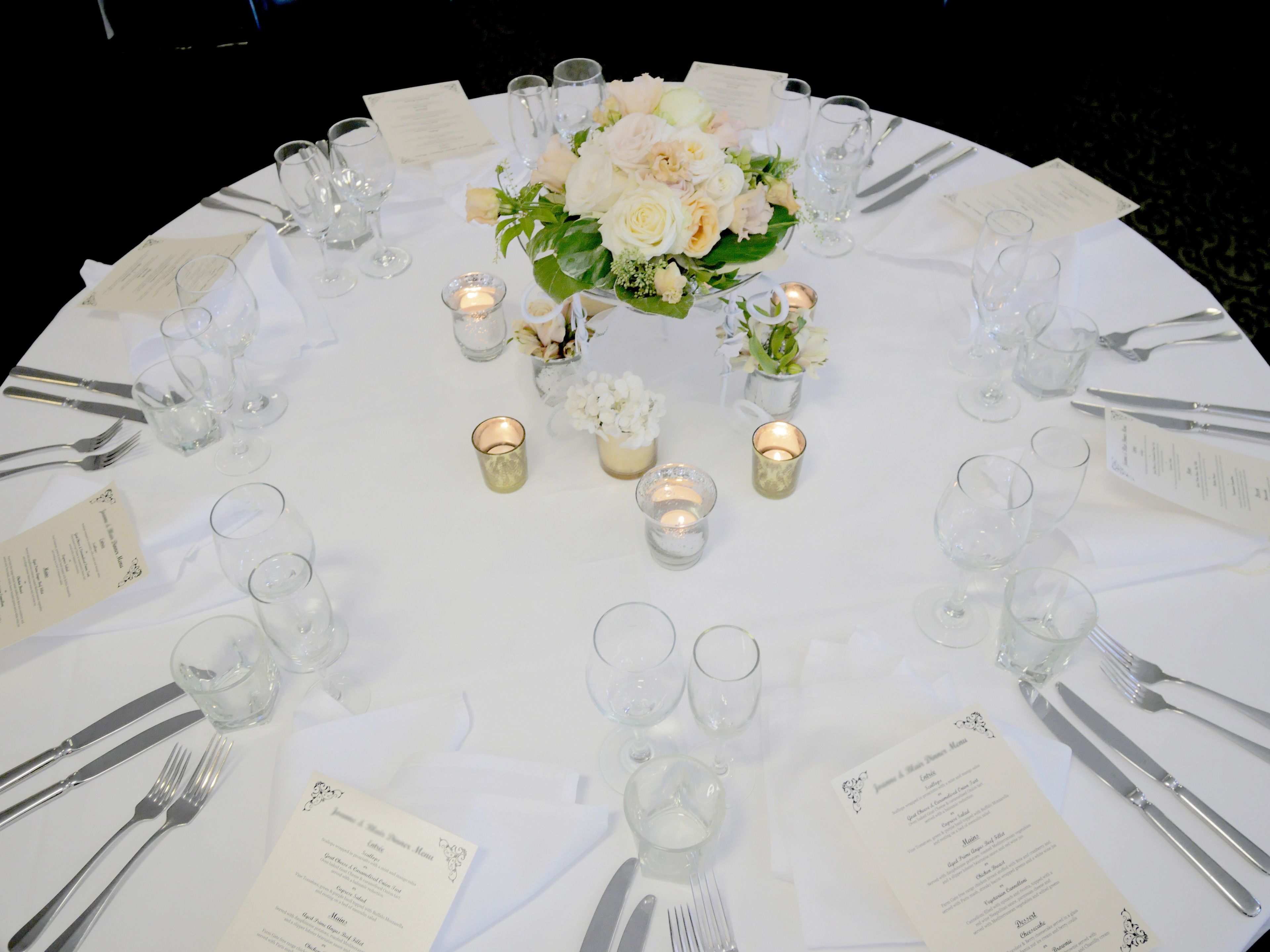A round table set for a formal event with white tablecloths, glassware, and floral centerpieces.