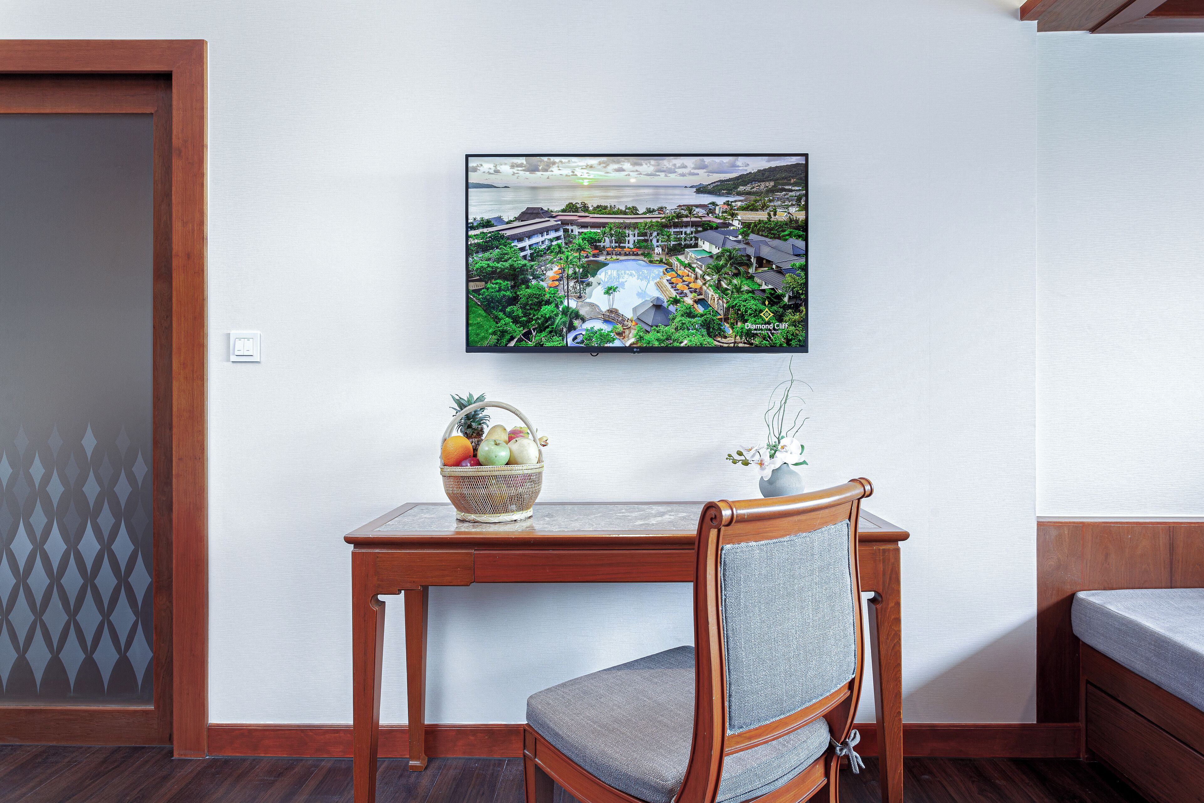 A hotel room with a wooden desk, a chair, a television mounted on the wall, and a small table with a fruit basket and a vase with flowers.