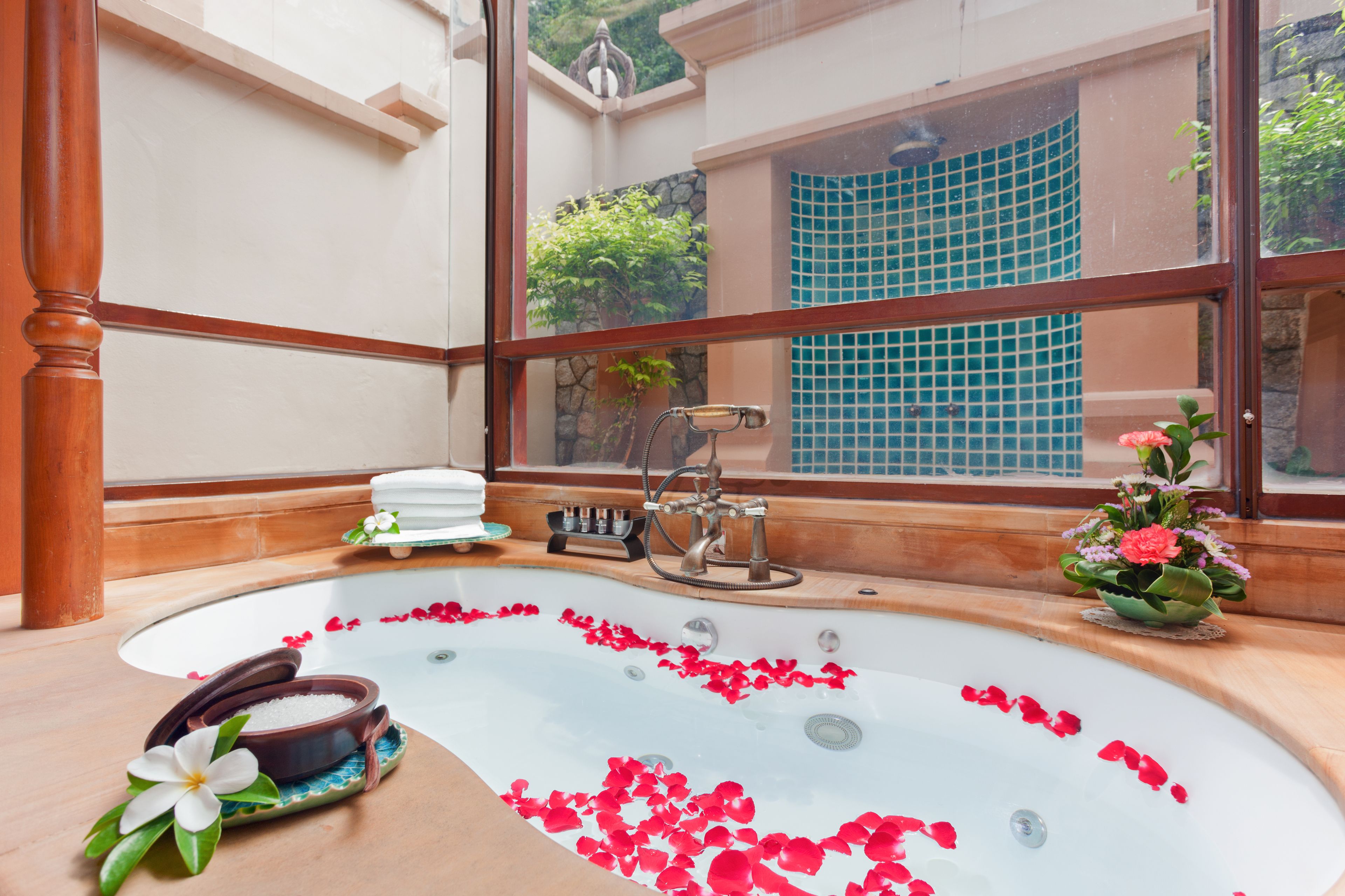 Une grande baignoire remplie d’eau et de pétales de rose, entourée de panneaux en bois et donnant sur une fenêtre avec vue sur la verdure.