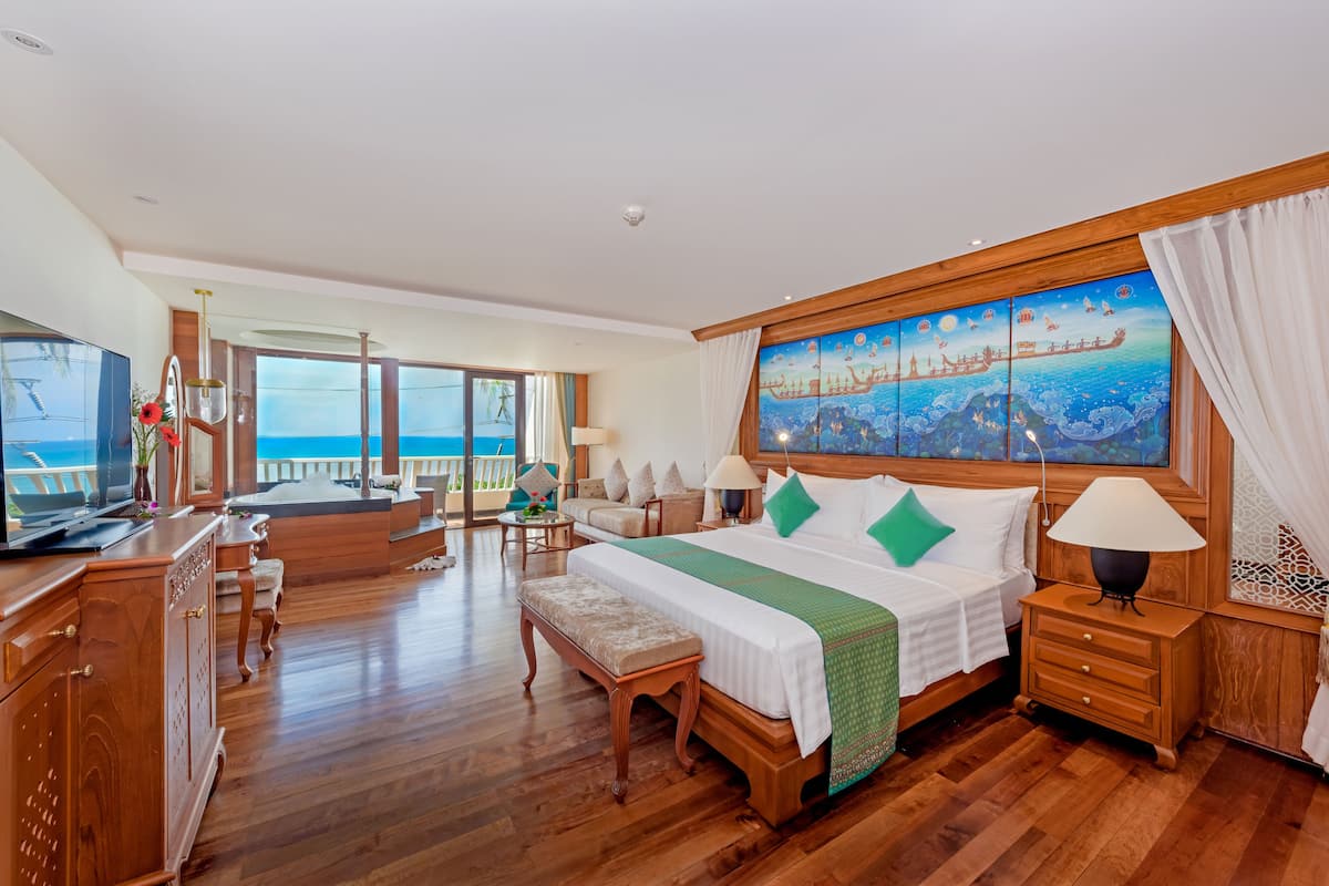 A spacious bedroom with a large bed, a television, a wooden dresser, and a balcony with a view of the ocean.