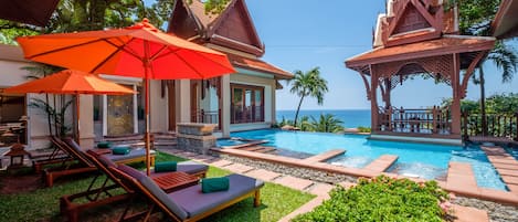 A pool area with lounge chairs and umbrellas, surrounded by tropical plants and a view of the ocean.