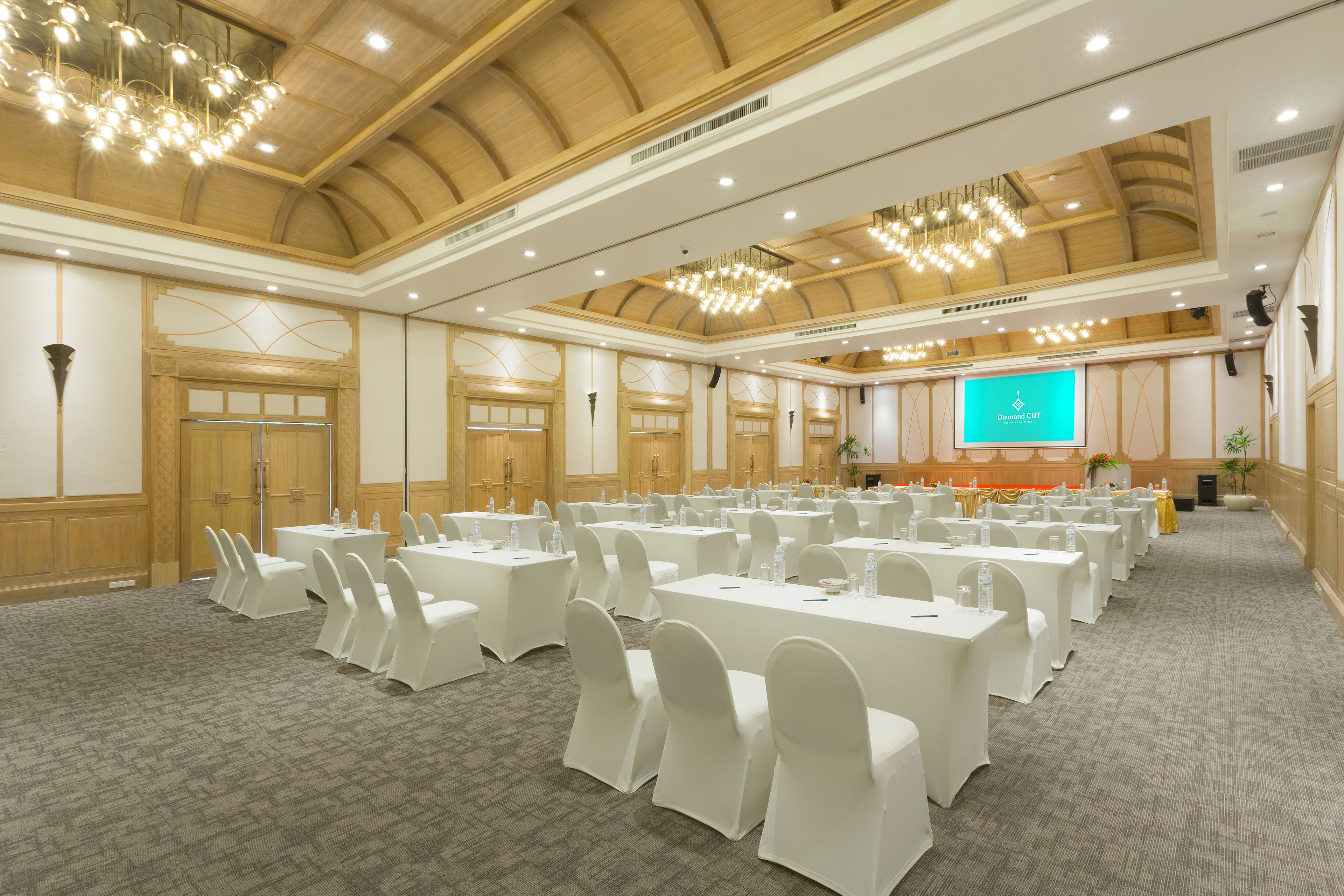 Uma sala de conferências ampla com fileiras de mesas e cadeiras brancas, teto de madeira e uma tela de projeção ao fundo.