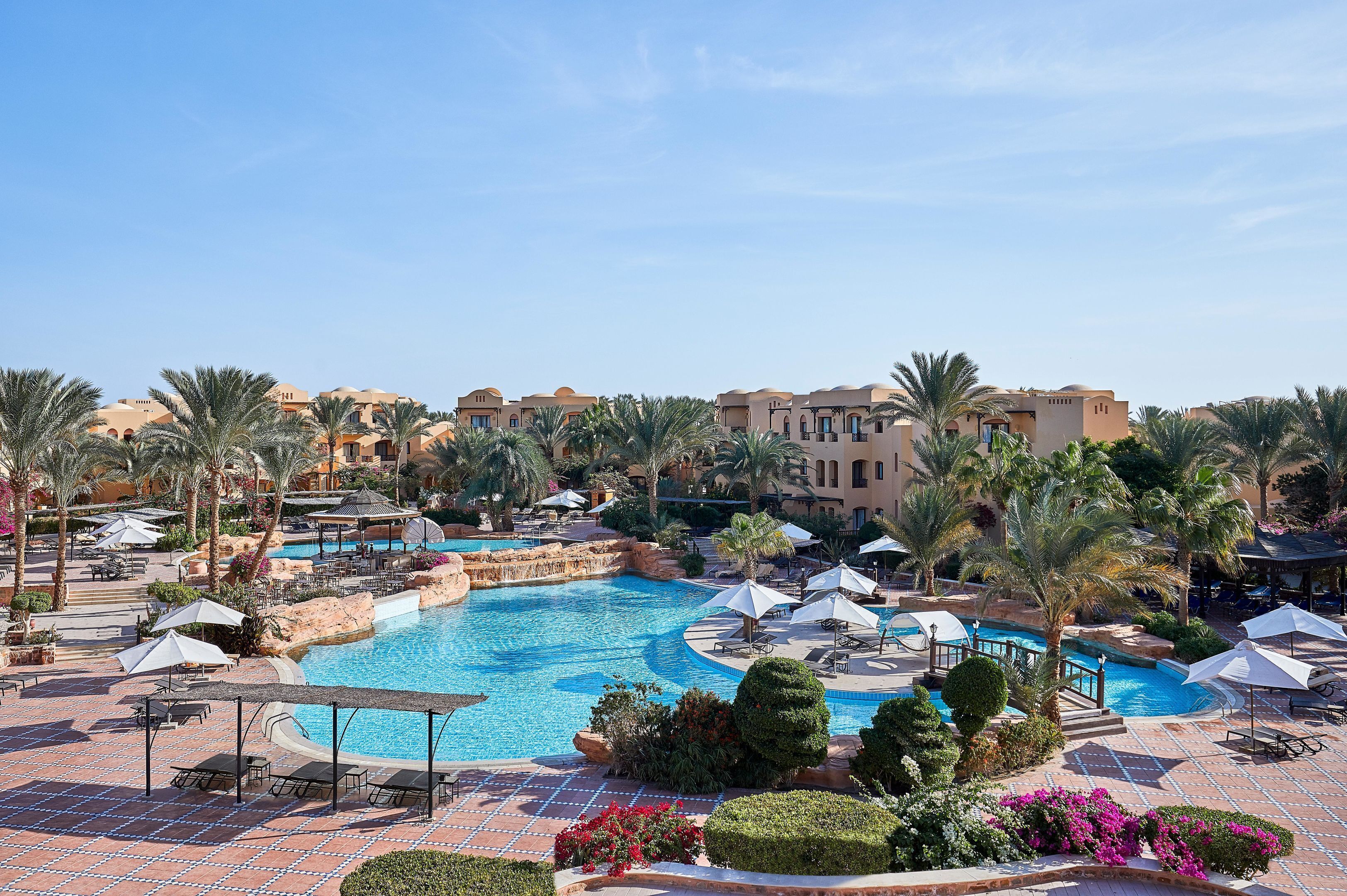 Ein Poolbereich eines Resorts mit Palmen, Liegestühlen und blauem Himmel.