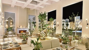 Un comedor interior bien iluminado con varias mesas dispuestas para comer, un área de estar central con un sofá y vistas del exterior durante la noche.