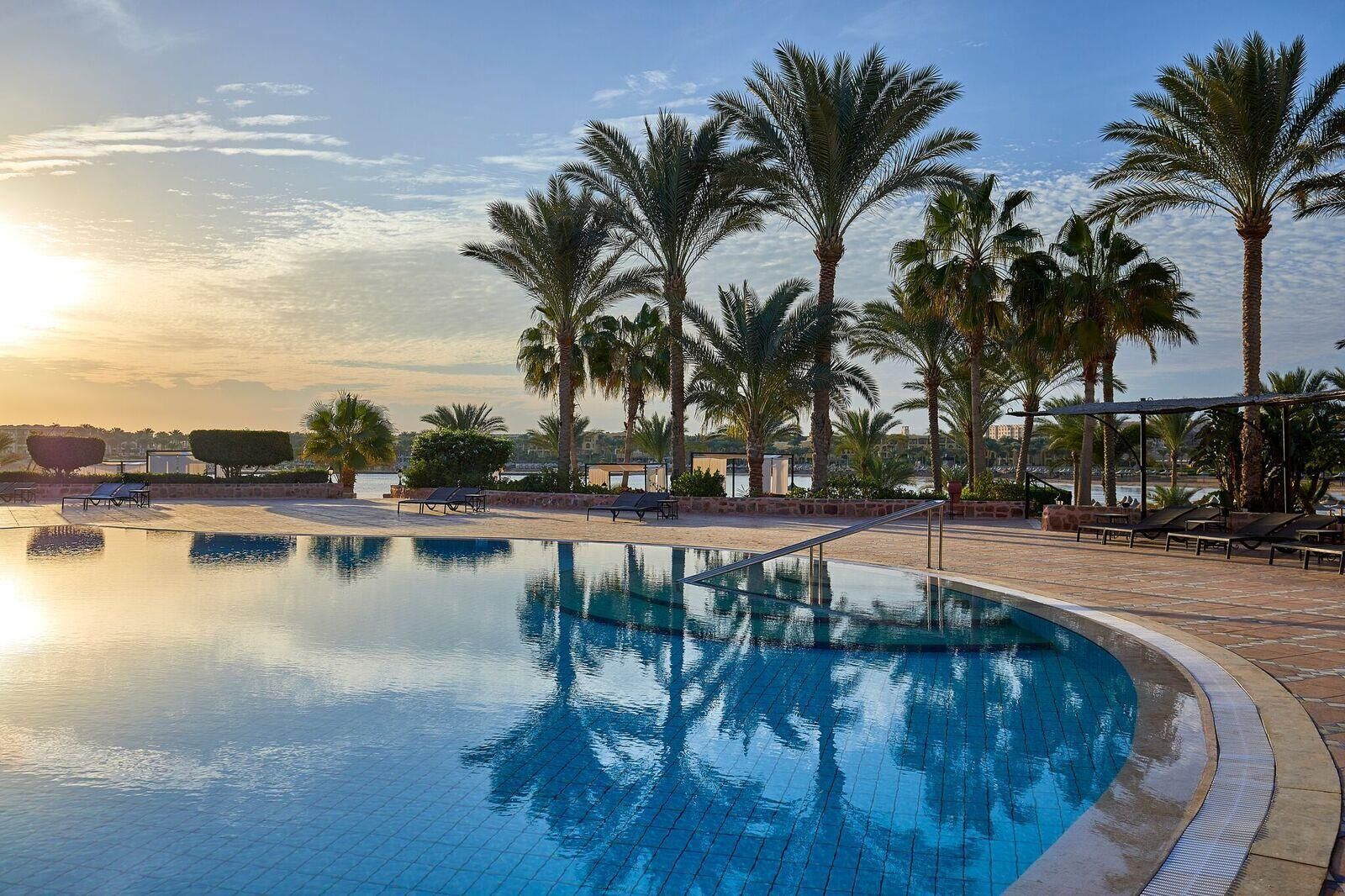 A swimming pool with lounge chairs and palm trees, surrounded by a stone deck and a view of the sea.