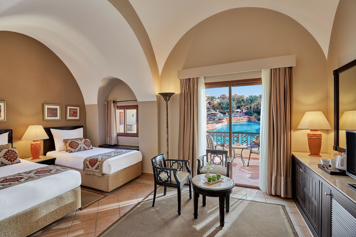 Habitación de hotel con dos camas, un área de estar con mesa y sillas, televisión y vista a una piscina.