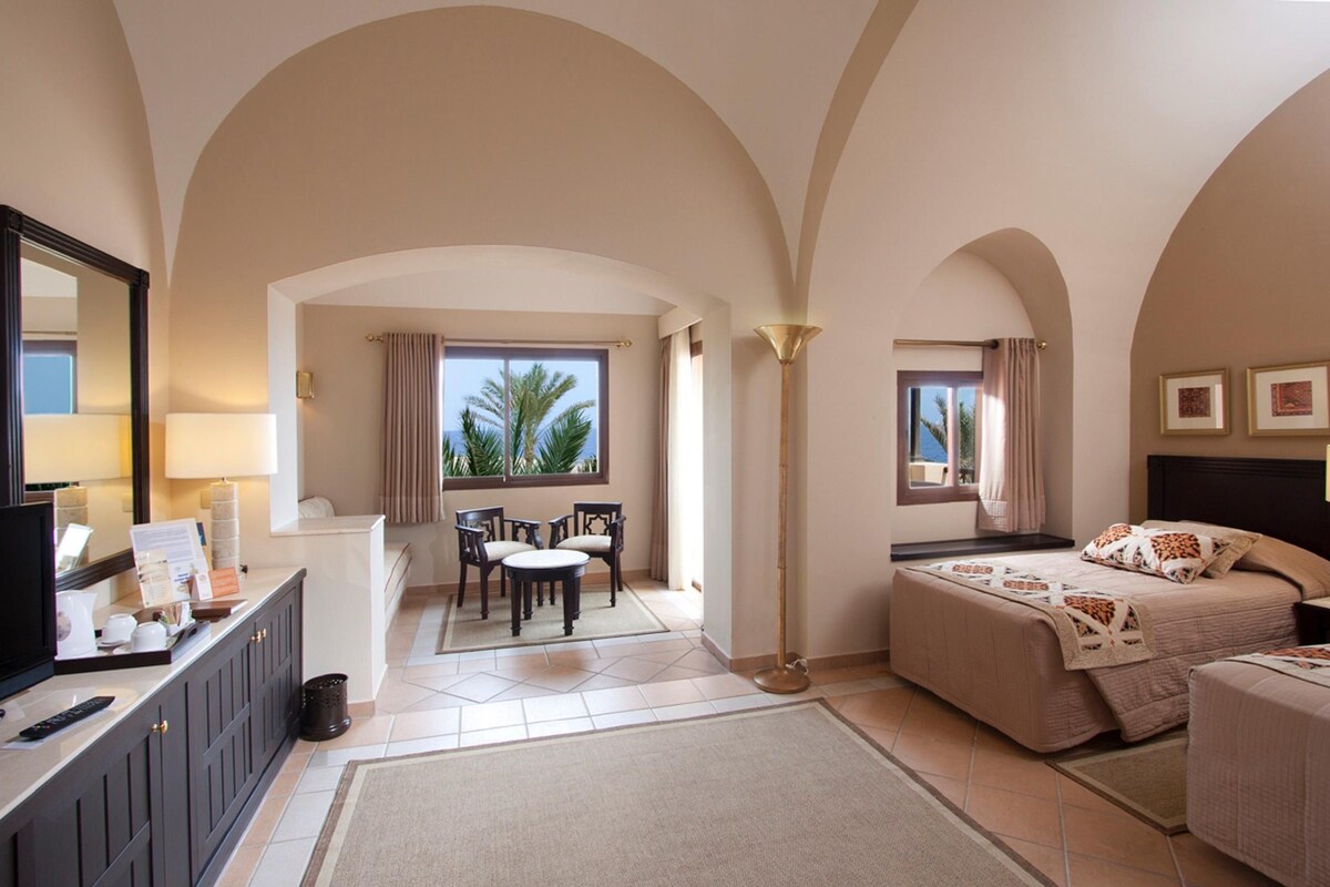 Habitación de hotel con cama, escritorio con televisor, una silla, lámpara y vistas a palmeras.
