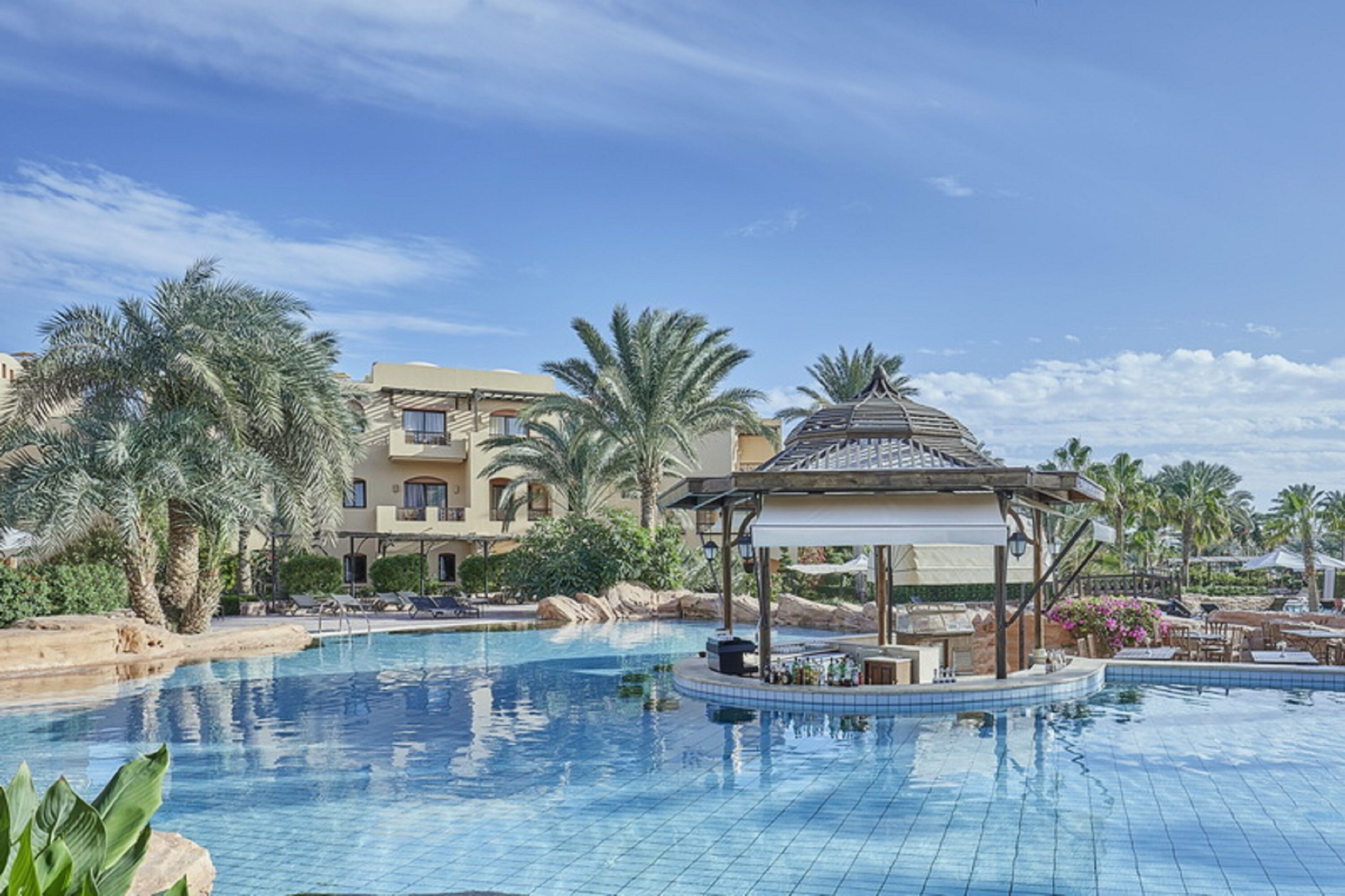 A swimming pool with a gazebo, palm trees, and a building with balconies in the background.