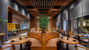 Un restaurante moderno con mesas y cabinas de madera, una planta en el centro y una zona de bar al fondo.