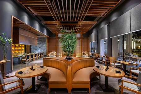 A modern restaurant with wooden tables and booths, a central plant, and a bar area in the background.