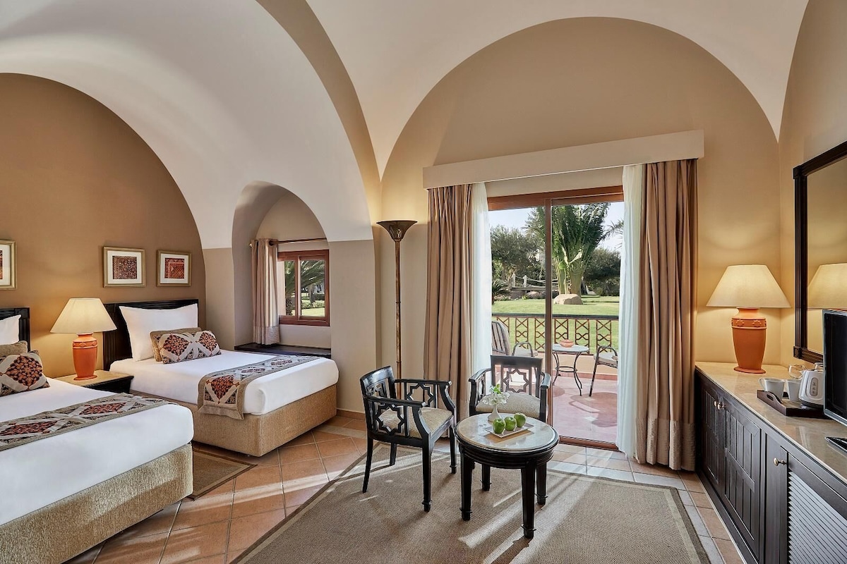 Habitación de hotel con dos camas, zona de estar con sillas y una mesa, balcón con vistas al jardín y un televisor en la cómoda.