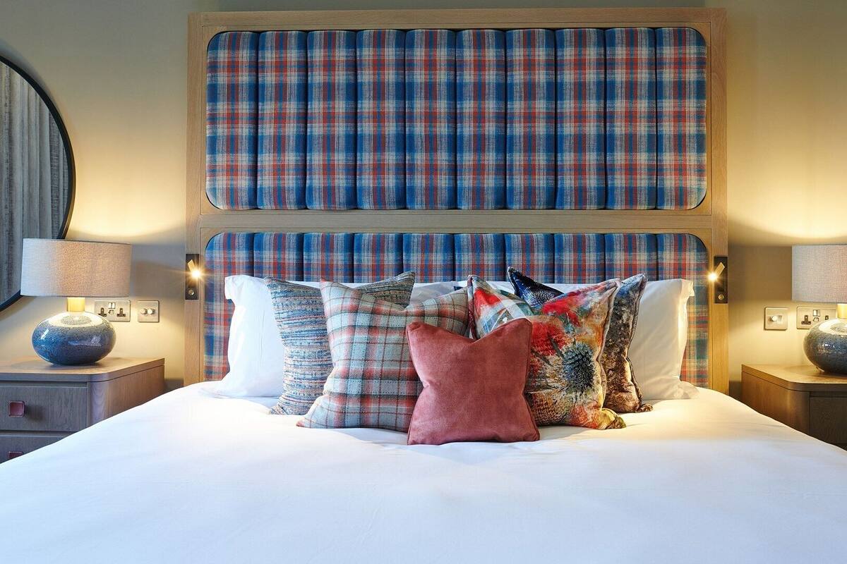 Una cama bien hecha con un tablero de cabecera escocés, dos mesitas de noche con lámparas y un espejo redondo en la pared.
