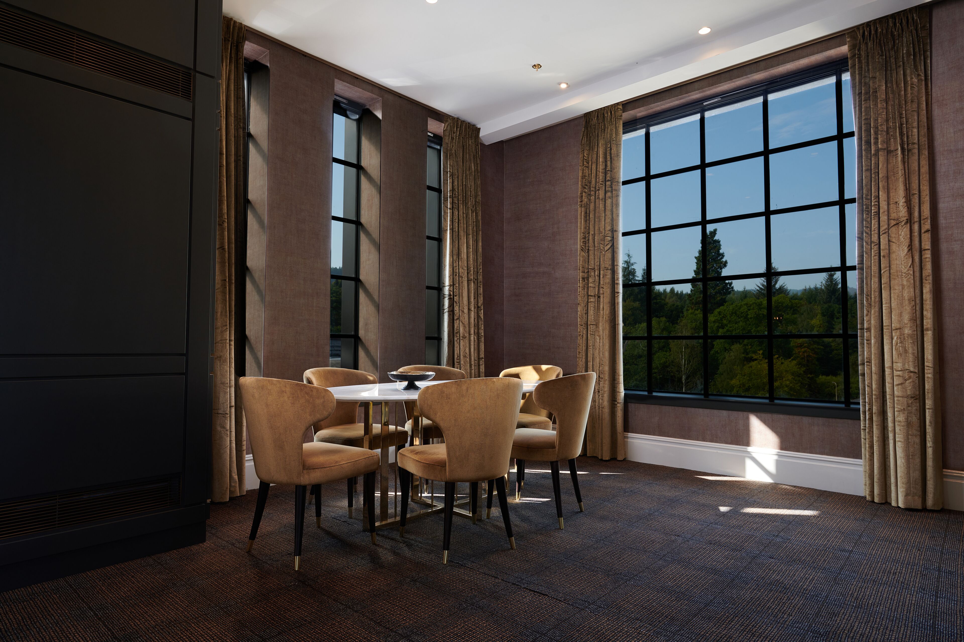 A dining area with a round table and six chairs, large windows with curtains, and a carpeted floor.