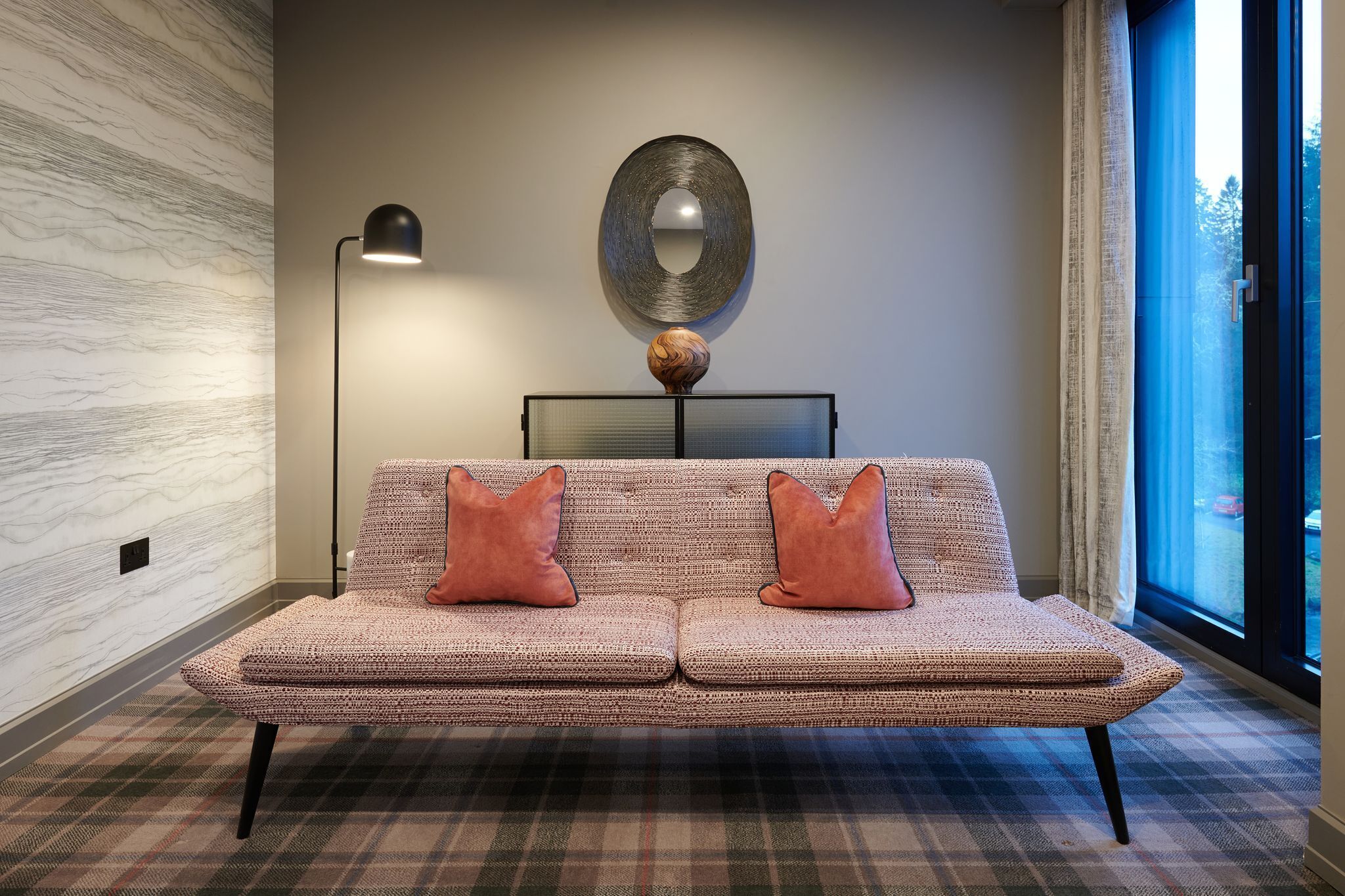 En moderne stue med en vævet sofa, to puder, en gulvlampe, et rundt spejl og et stort vindue med gardiner.