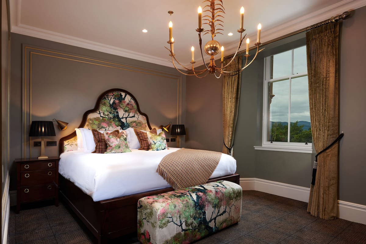 Una cama bien hecha con un reposapiés estampado con motivos florales, una araña y una ventana con cortinas.