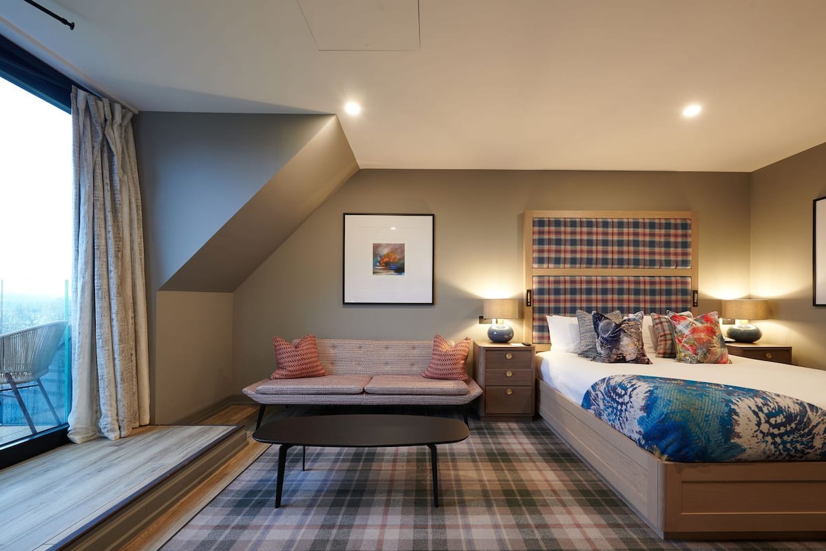 Un dormitorio con cama, sofá, mesita de noche, lámpara y ventana con cortinas.