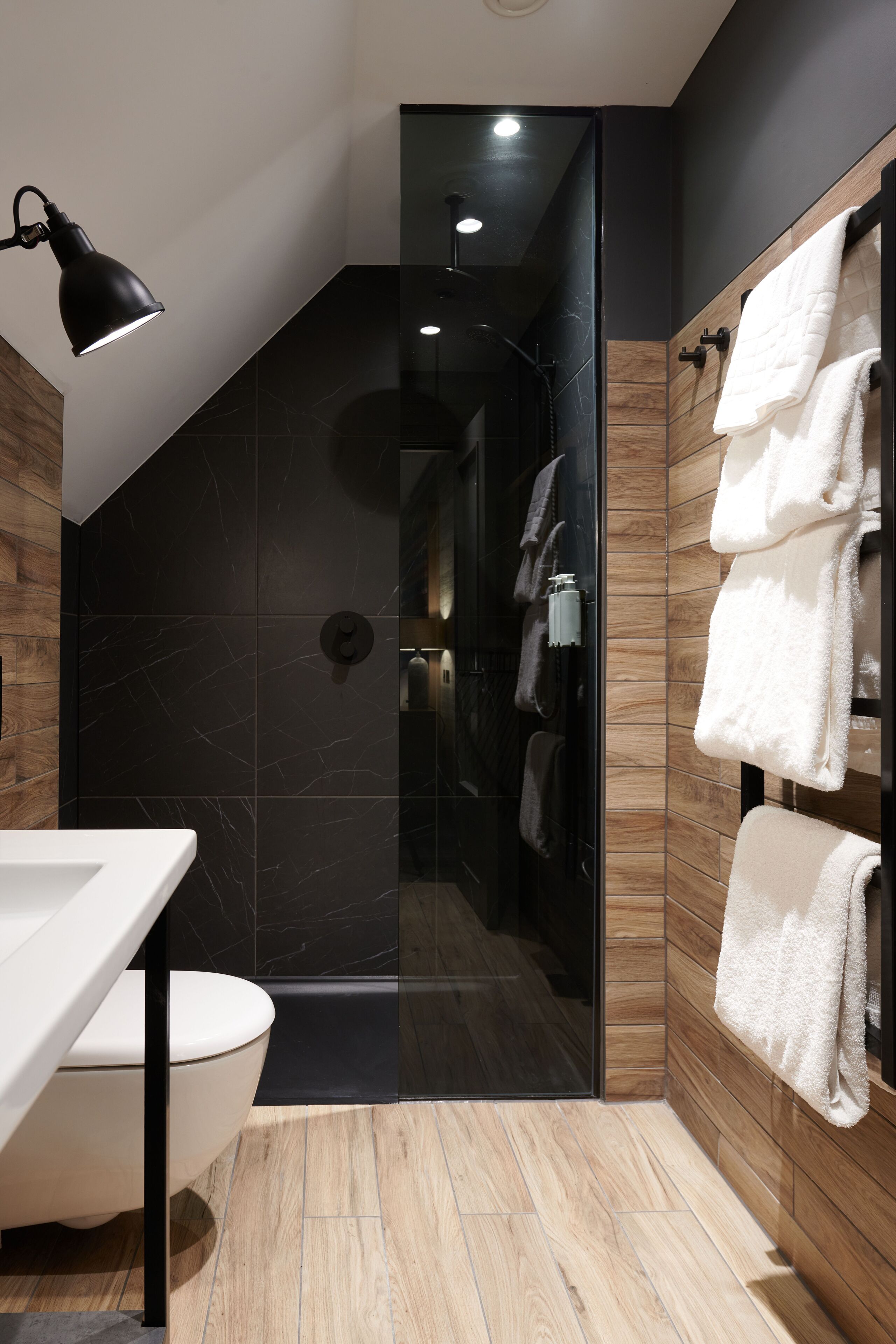 Un baño moderno con un lavamanos blanco, paredes de mármol negro, un mueble de tocador de madera y una cabina de ducha de vidrio.