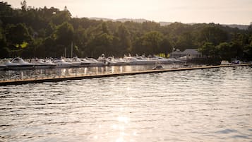 A marina with multiple boats docked along a wooden pier, surrounded by dense forest and hills in the background.