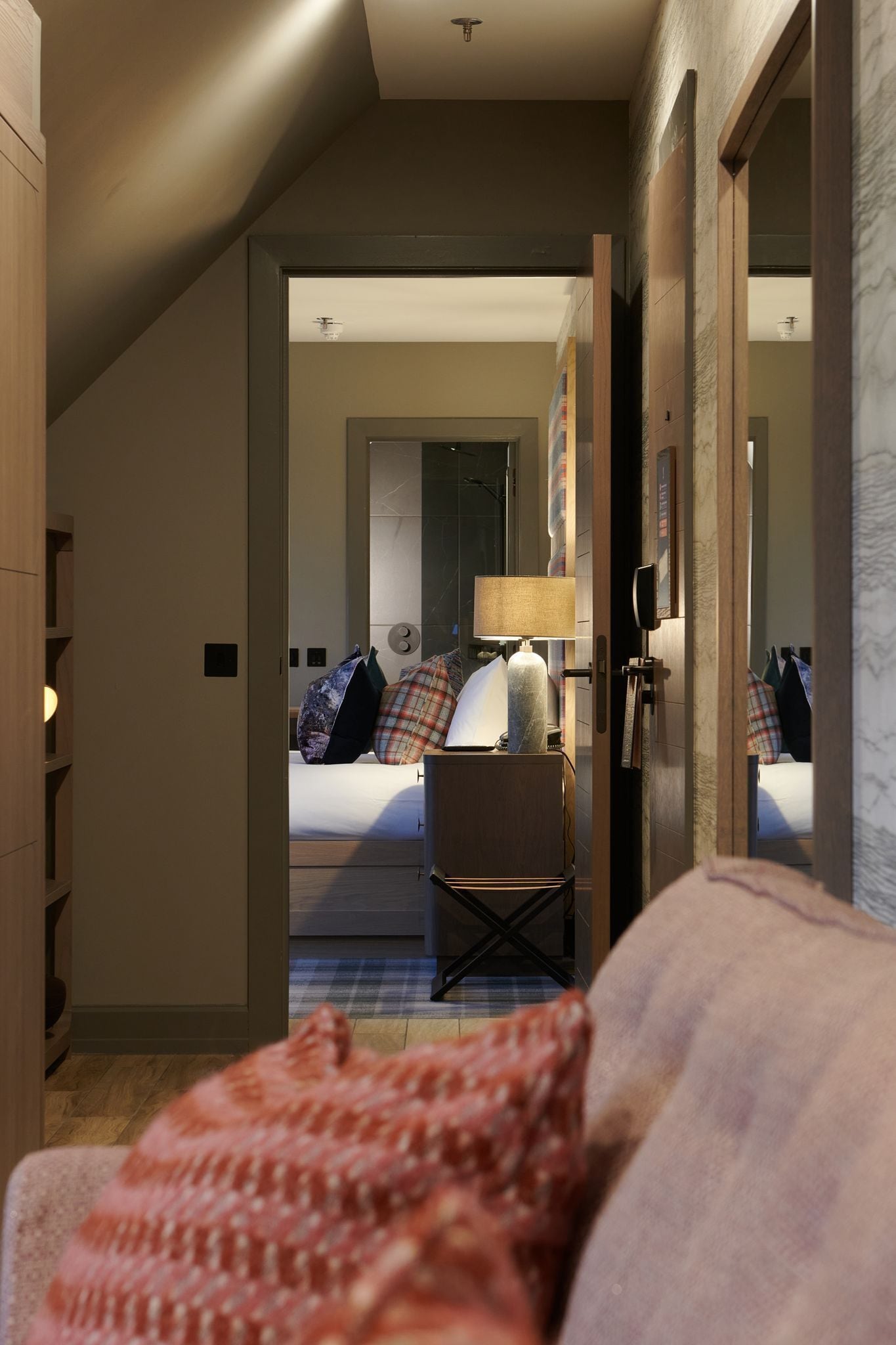 A hotel room with a bed, a desk, and a chair. There is a mirror on the wall, and a door leading to another room.