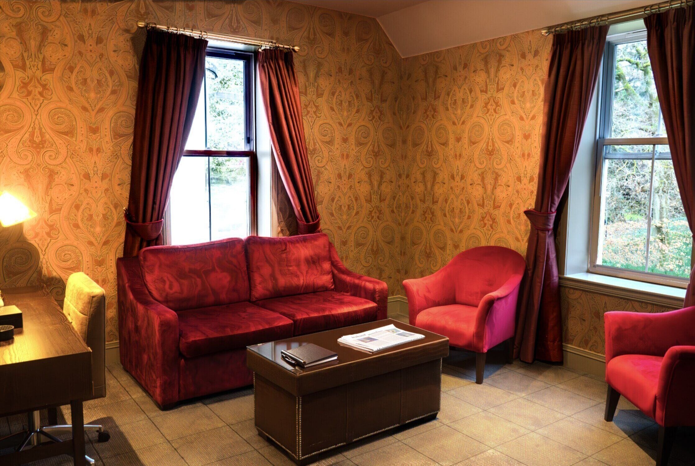 A room with a red sofa, a red armchair, and a wooden coffee table. There is a window with curtains and a lamp on a table.