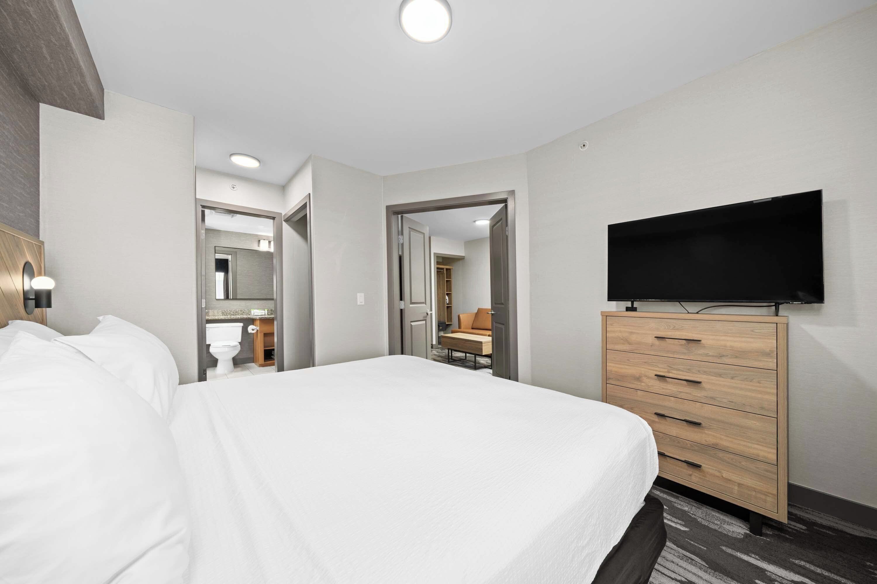 Ett modernt hotellrum med en stor säng, en byrå i trä, en platt-TV och ett badrum som syns genom en öppen dörr.