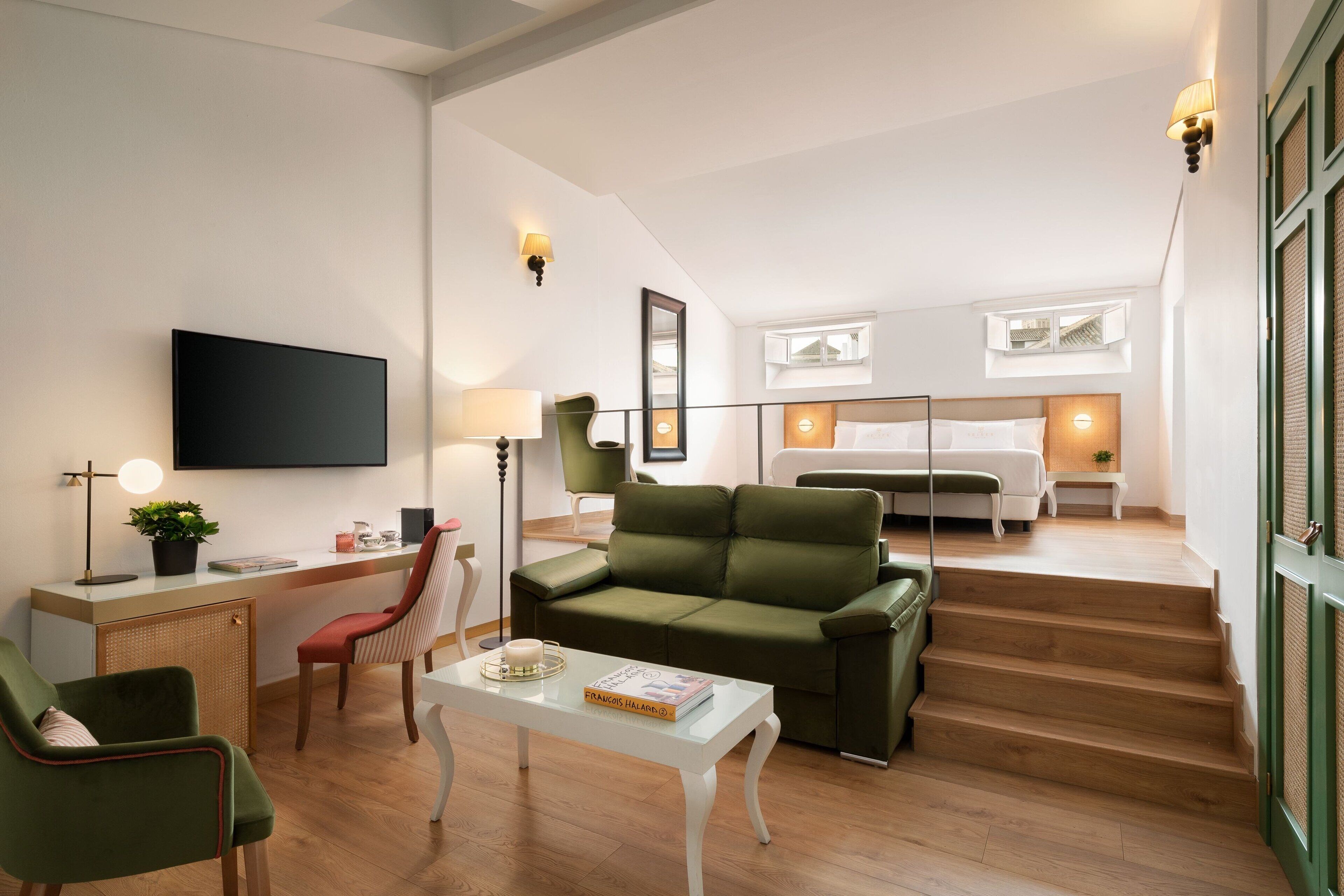 A modern living room with a sofa, a coffee table, a desk, and a TV mounted on the wall. There is a staircase leading to a bedroom with a bed and a window.