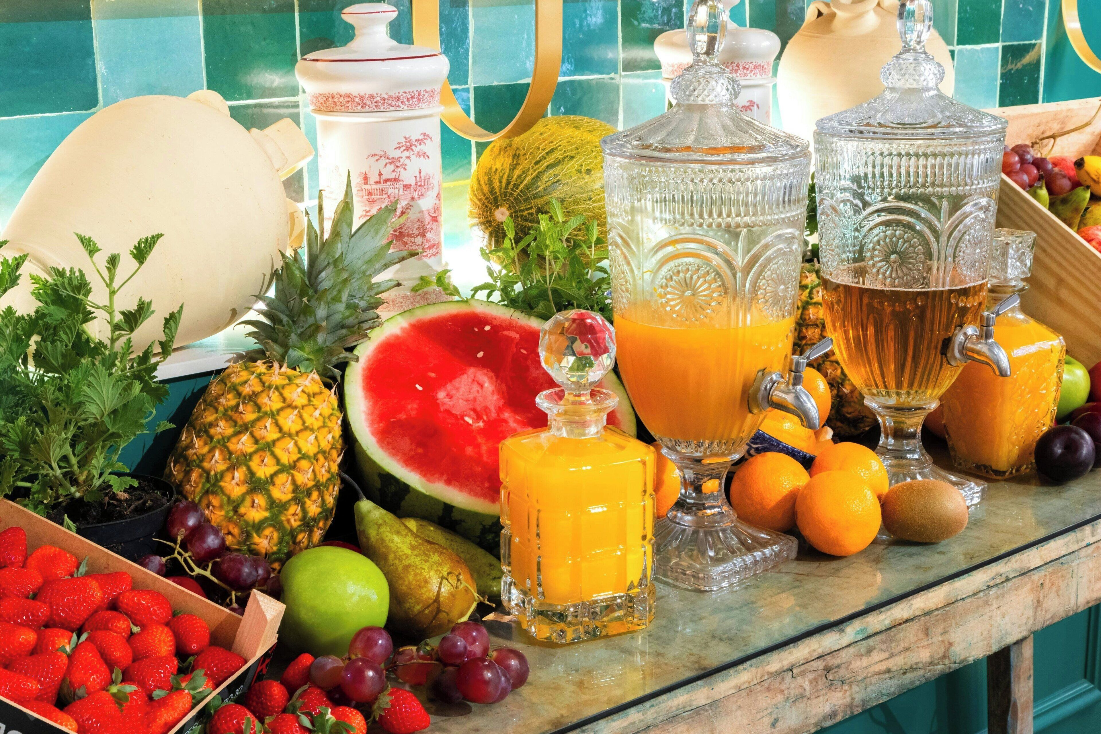 A kitchen counter with various fruits, a pitcher of orange juice, and a bottle of honey.