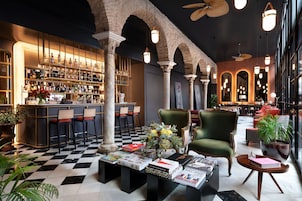 A bar area with a checkered floor, bar stools, and a bar counter with bottles. There are chairs and a table with magazines and books.