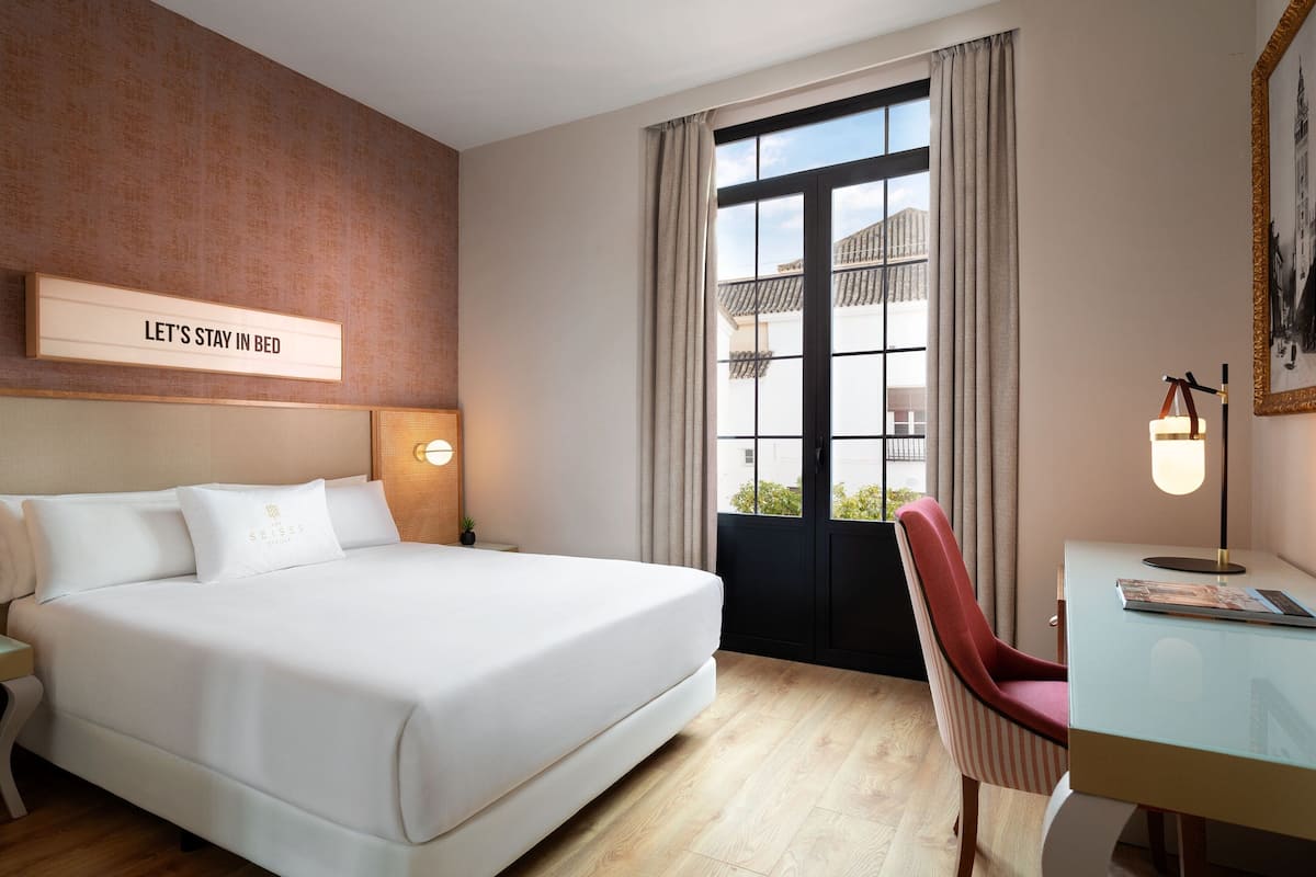Hotellihuone, jossa on suuri sänky, työpöytä tuolilla, ikkuna verhoilla ja seinällä kyltti.