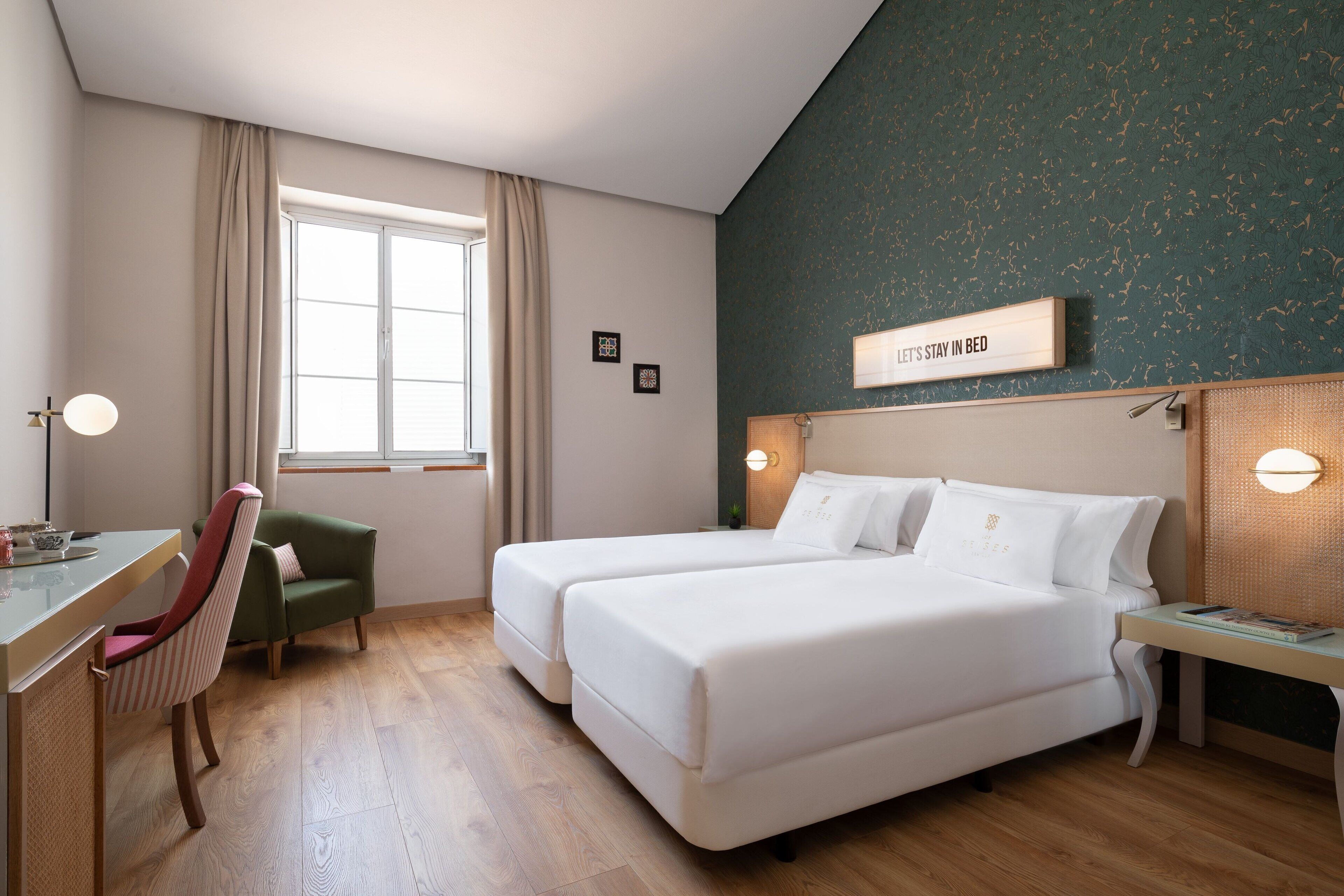 A hotel room with two beds, a desk, a chair, a window with curtains, and a sign on the wall.