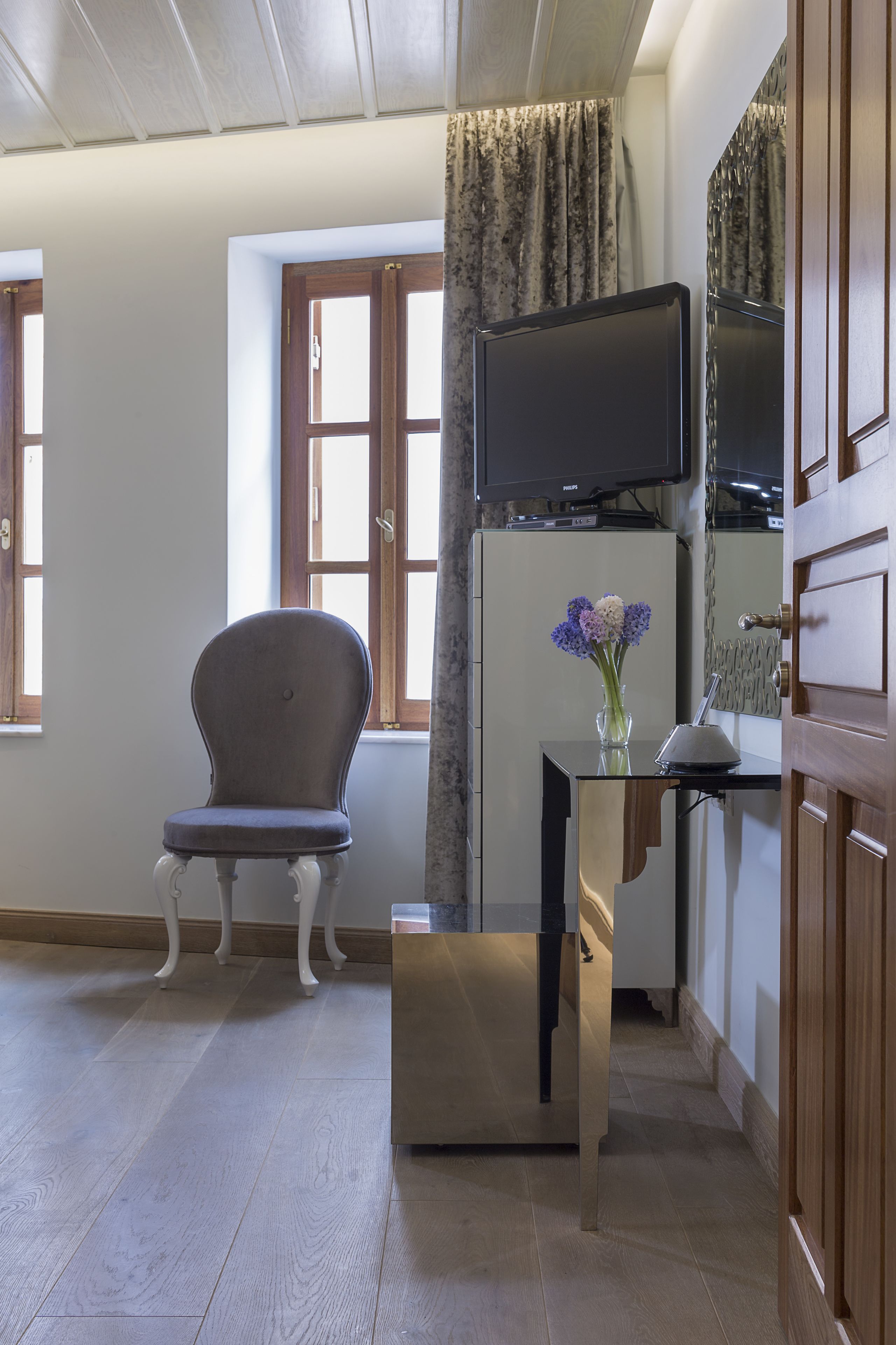 Una habitación con piso de madera, una silla, una mesita con un jarrón de flores, un televisor de pantalla plana montado en un mueble y una ventana con cortinas.