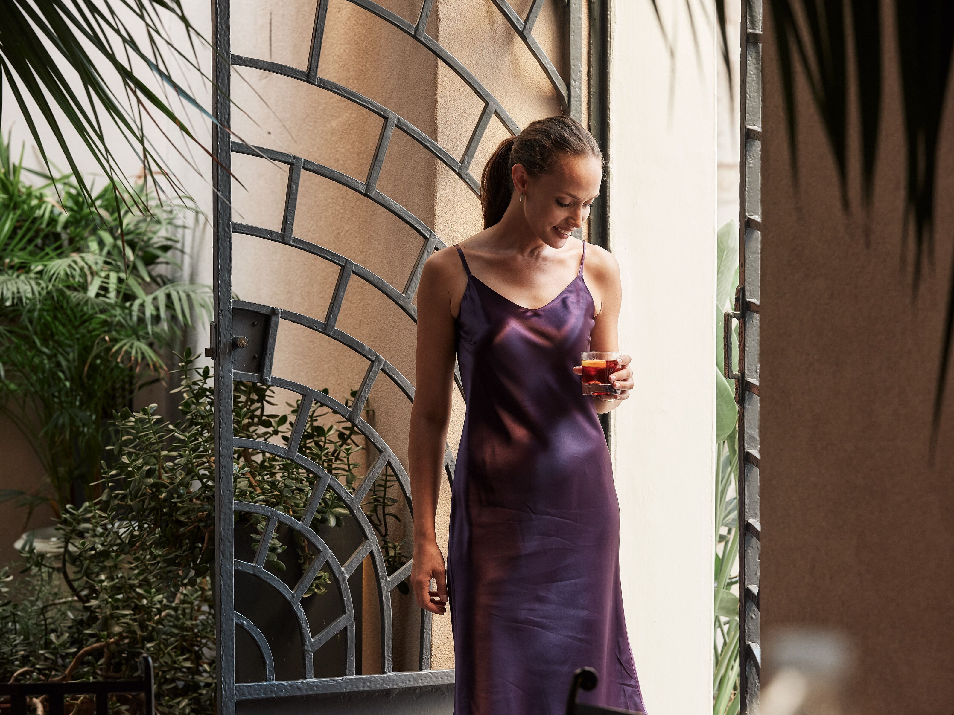 A person in a purple dress standing near a metal gate with plants in the background.