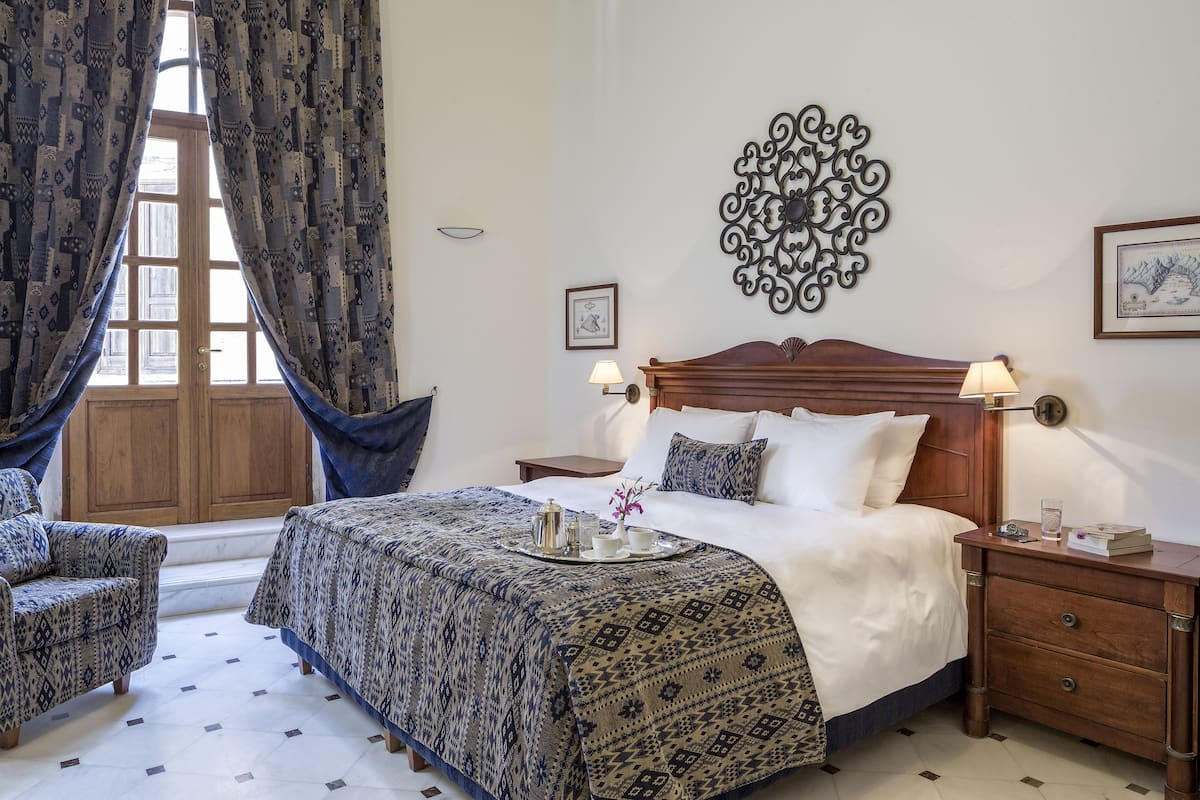 Ein Hotelzimmer mit einem großen Bett, zwei Nachttischen, einem Holzbeistelltisch, einem Sessel sowie einer Auflage mit Teeservice und gemusterten Vorhängen.