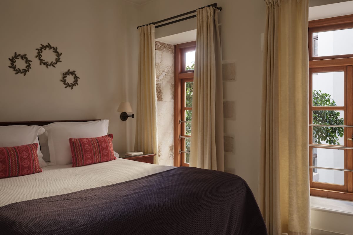 Ein Schlafzimmer mit einem Bett, zwei Kissen, einem Nachttisch, einer Wandlampe und einem Fenster mit Vorhängen.