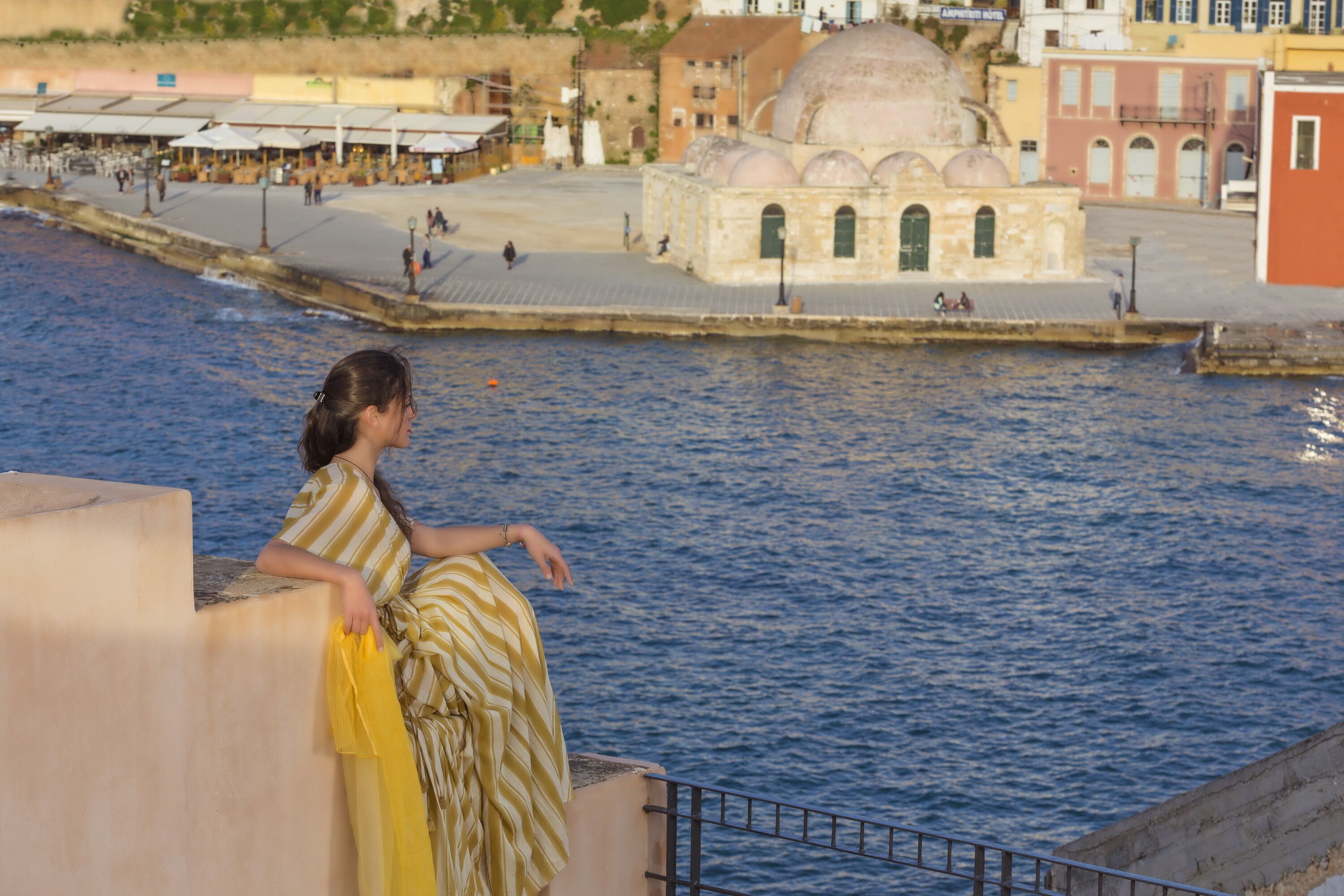 A woman in a striped dress leaning on a railing by a waterfront, overlooking a cityscape with buildings and a domed structure.