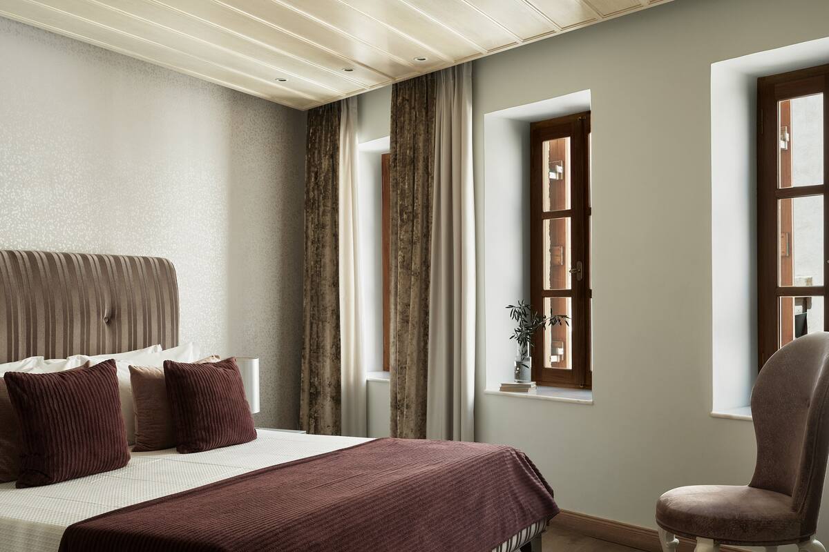 Ein Schlafzimmer mit einem Bett, einem Sessel, einem Fenster mit Vorhängen und einer Pflanze auf dem Fensterbrett.