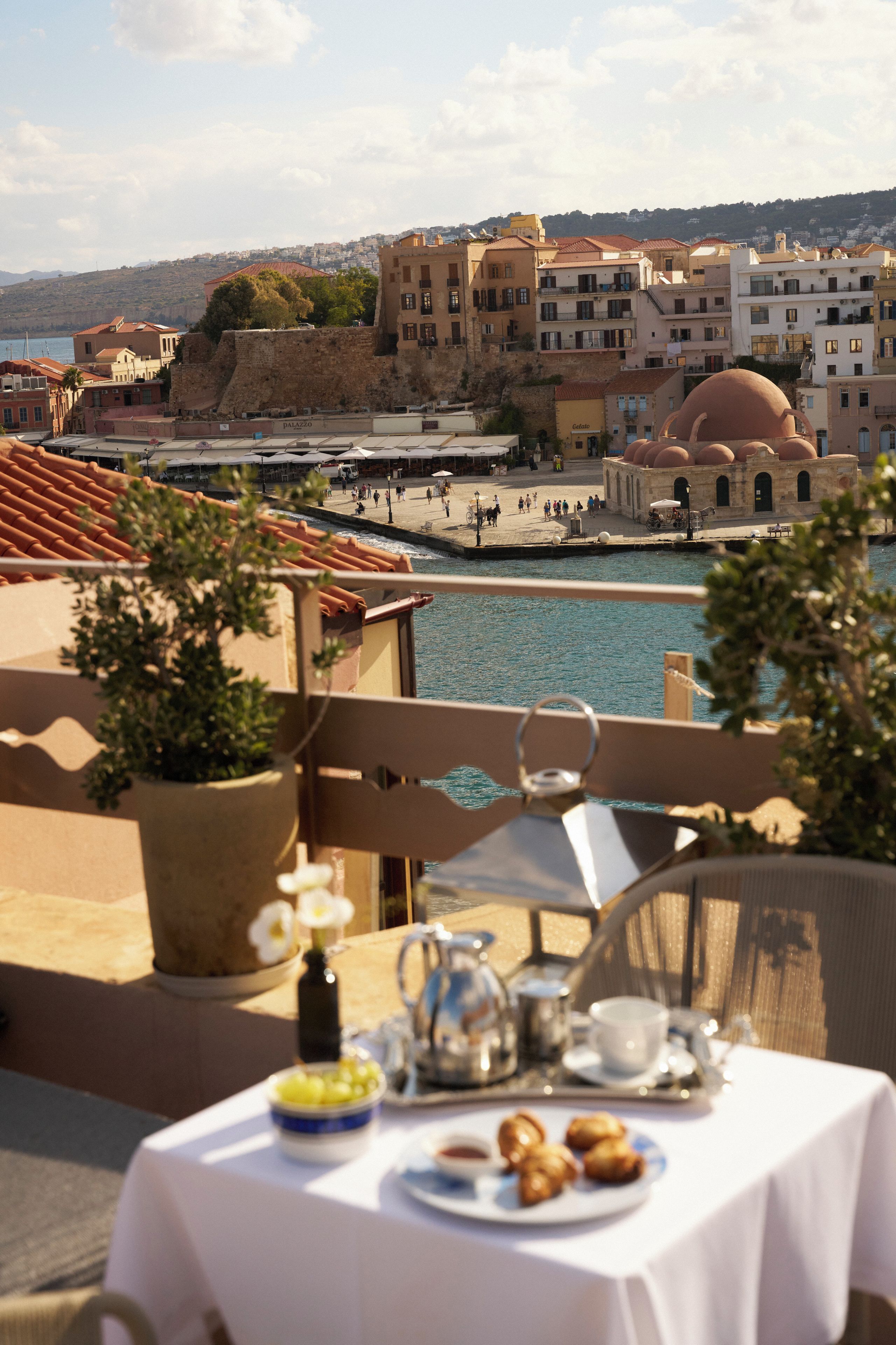 Una mesa dispuesta con alimentos para el desayuno en un balcón con vistas a un pueblo costero y edificios históricos.