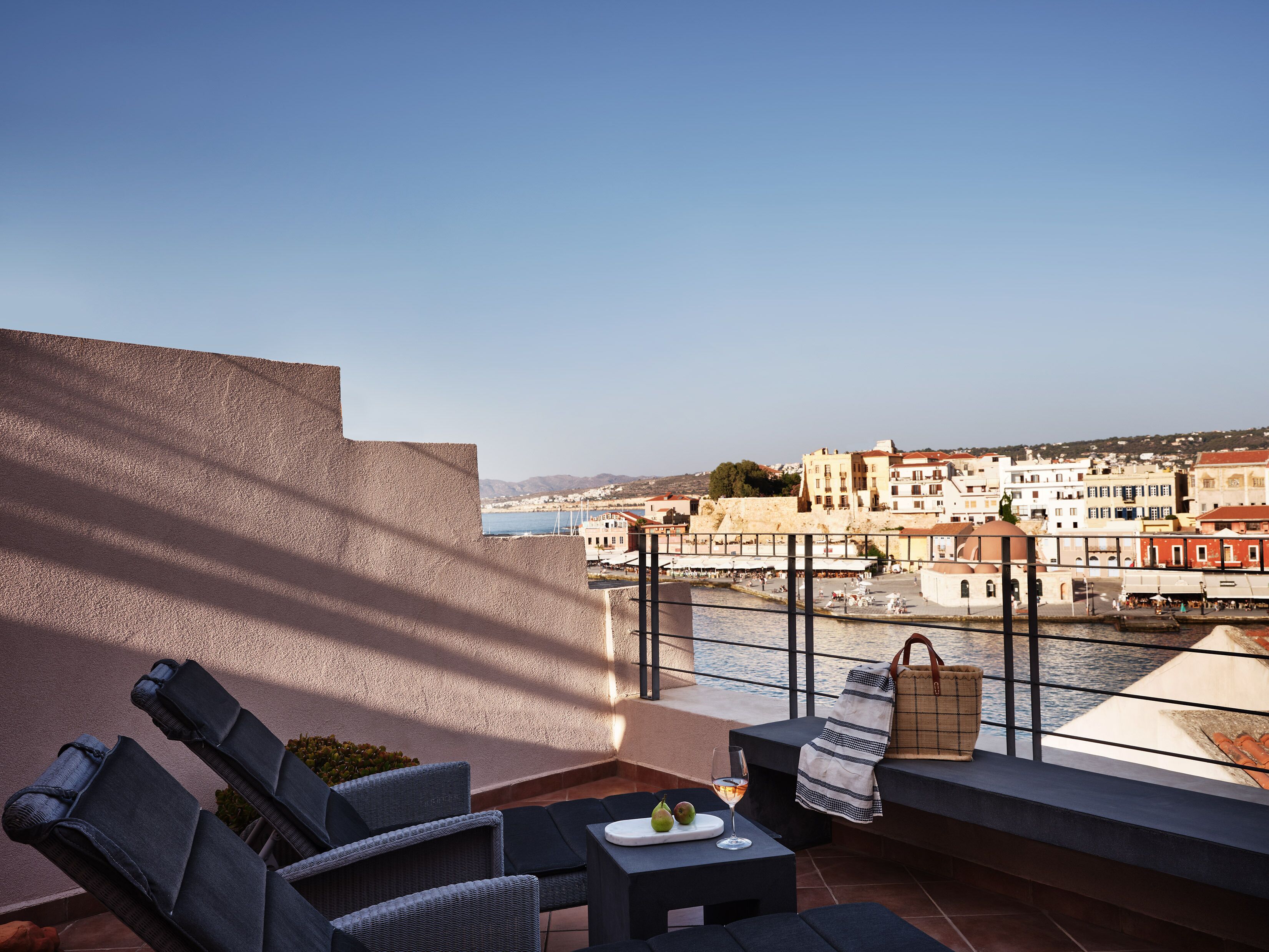 A terrace with wicker furniture, a table with a glass of wine and fruit, and a view of a coastal town with buildings and a river.