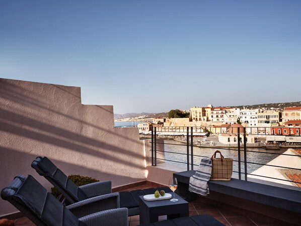 A terrace with wicker furniture, a table with a glass of wine and fruit, and a view of a coastal town with buildings and a river.