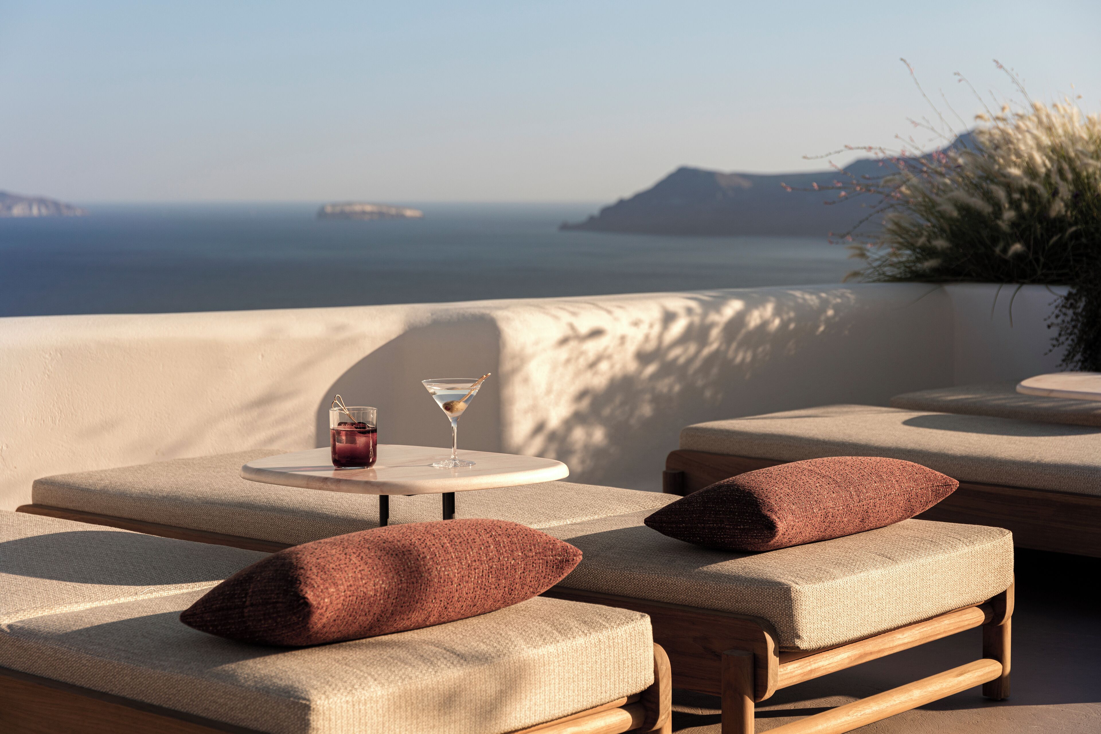 Un espacio exterior iluminado por el sol con sillas acolchadas y una mesa, con vistas a la costa y formas terrestres en la distancia.