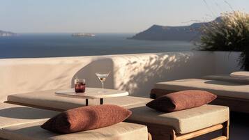 Un espacio exterior iluminado por el sol con sillas acolchadas y una mesa, con vistas a la costa y formas terrestres en la distancia.