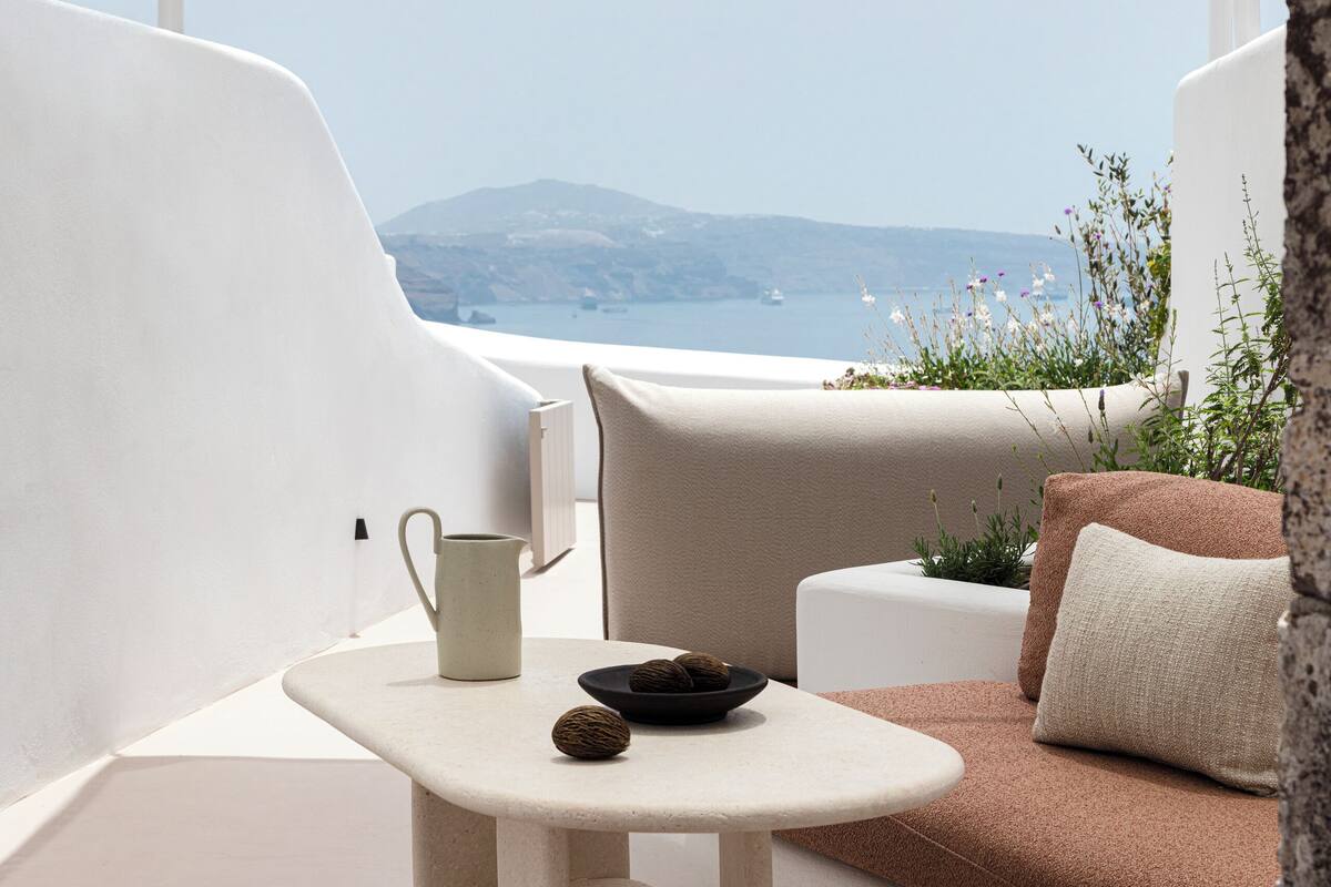A modern outdoor seating area with a table and chairs, a cushioned sofa, and a view of the sea and distant hills.