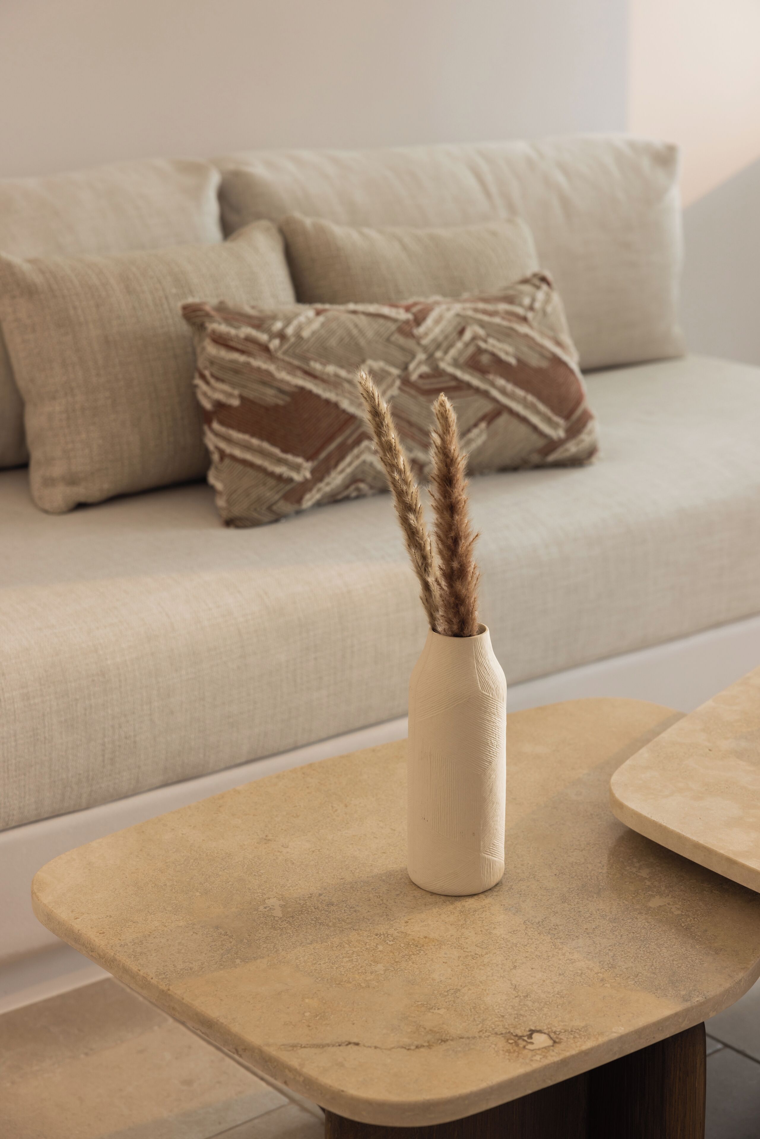 Un sofá color beige con cojines estampados y un jarrón con plumas de pluma en una mesa de centro texturizada.