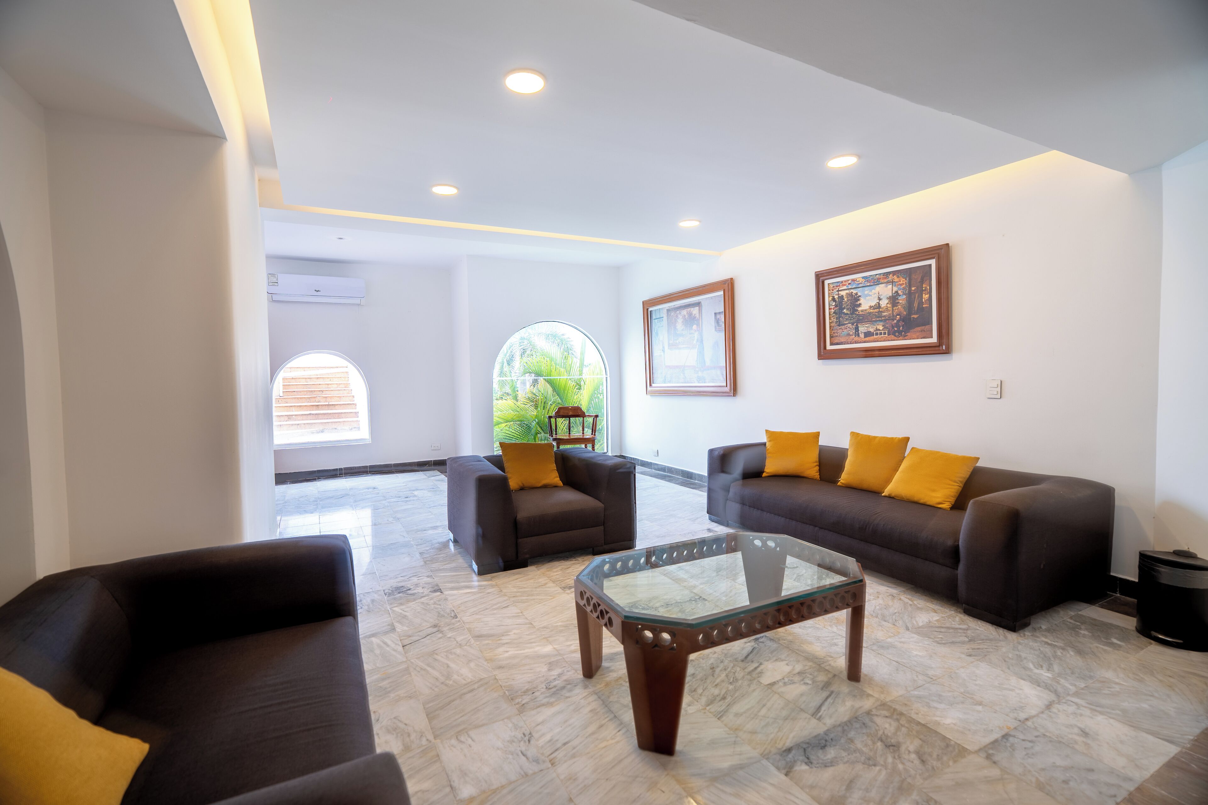 A spacious living area with two sofas and a glass coffee table, a large window with a view of greenery, and a framed picture on the wall.