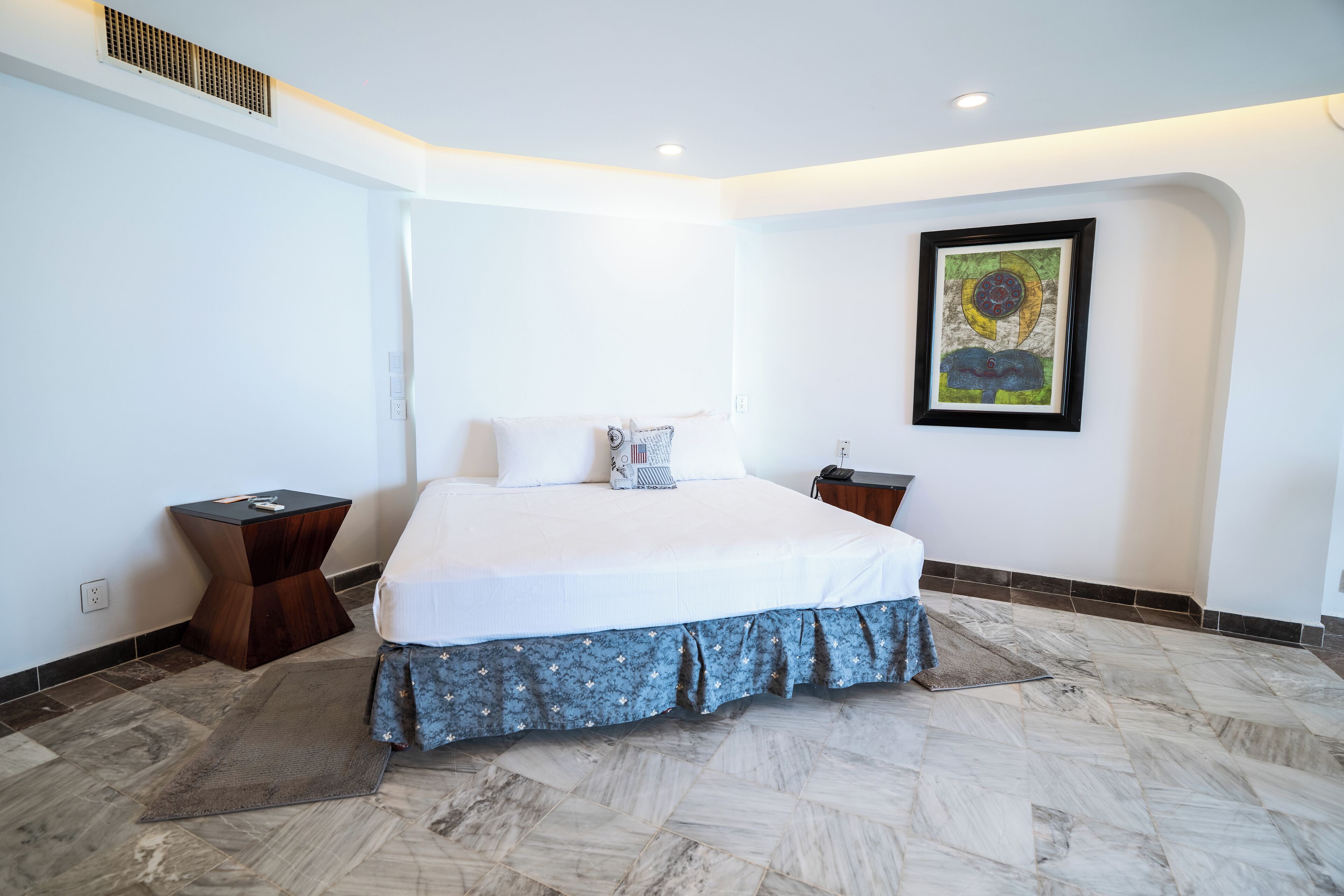Habitación de hotel moderna con una cama grande, una mesita auxiliar pequeña, una obra de arte en la pared y una alfombra estampada.