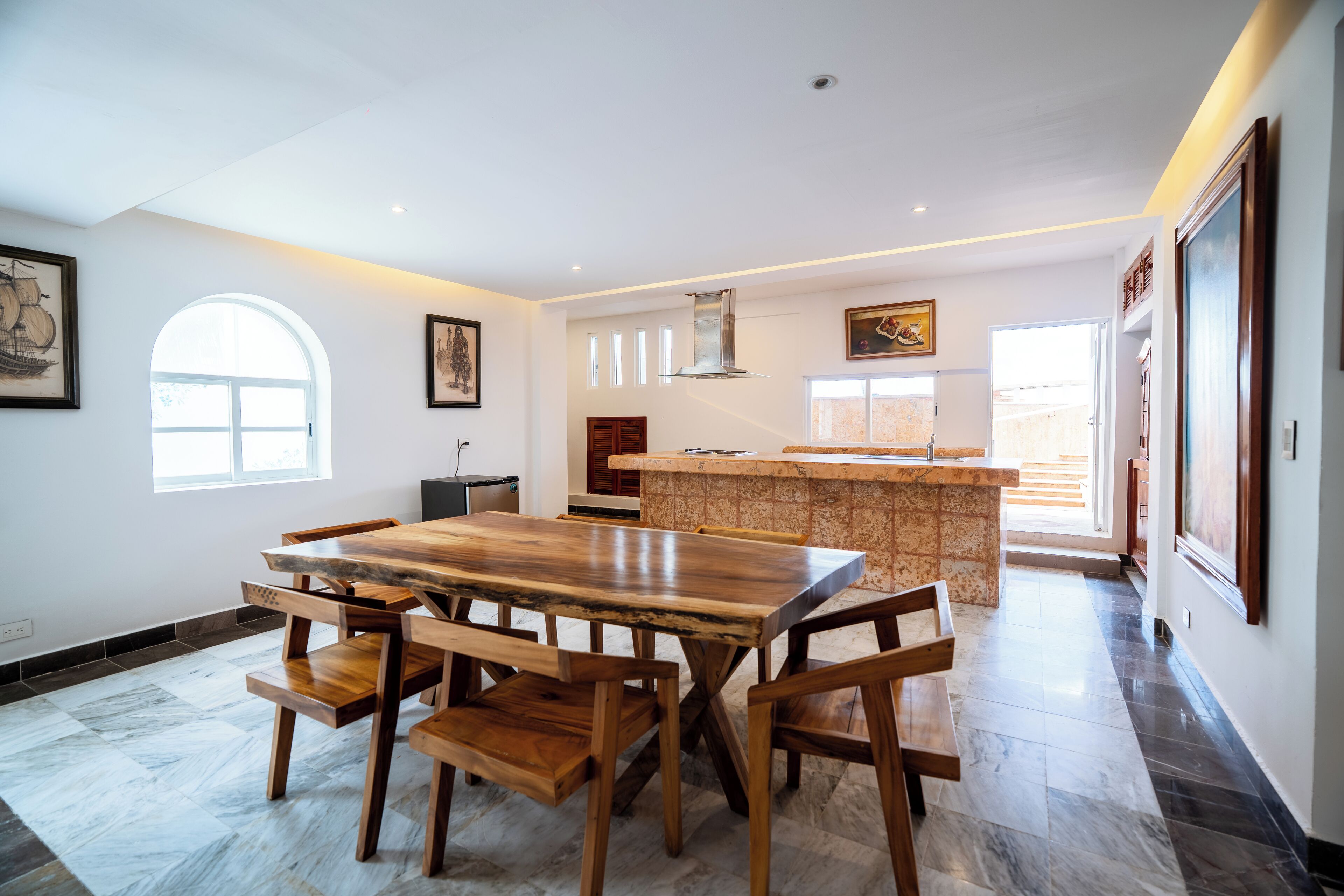 A spacious dining area with a large wooden table and chairs, a kitchen with a stone island, and a view of the outdoors through a large window.