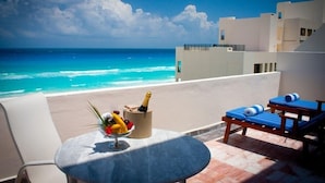 Un balcón con una mesa dispuesta con frutas y una botella, con vista a una playa y al océano.