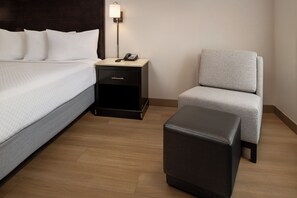 Een hotelkamer met een bed, een nachtkastje met een telefoon, een stoel en een klein voetenbankje.